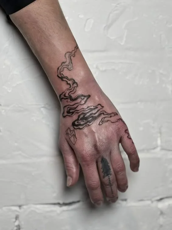 A hand with tattoos, including a smoking pipe with smoke rising from it, a small geometric shape, and a pine tree on one of the fingers, against a white brick wall background.