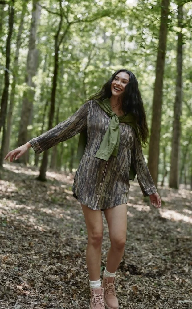 brunette model standing in the woods wearing a camo dress and backwards hat