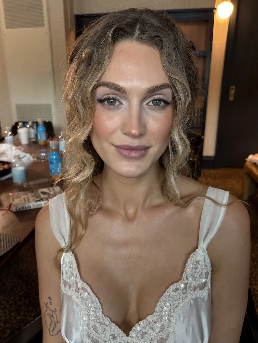 Close-up of a young woman with wavy blonde hair and blue eyes, smiling softly at the camera, wearing a white lace-trimmed sleeveless top, with a tattoo visible on her upper arm, in a room with a table and various items in the background.