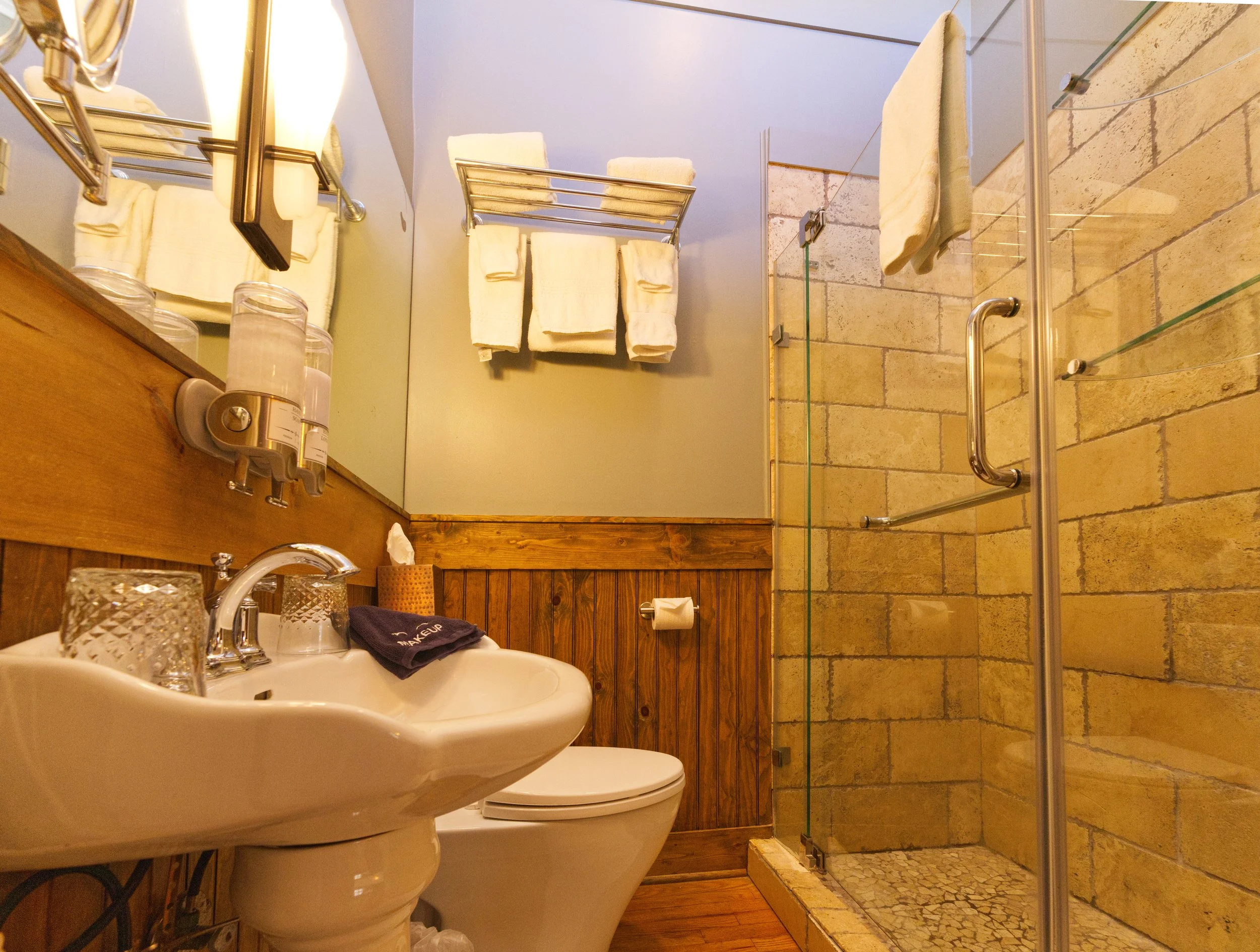 A small bathroom with wood and brick decor, featuring a toilet, a sink with a black towel, and a glass-enclosed shower with towels hanging on metal racks.