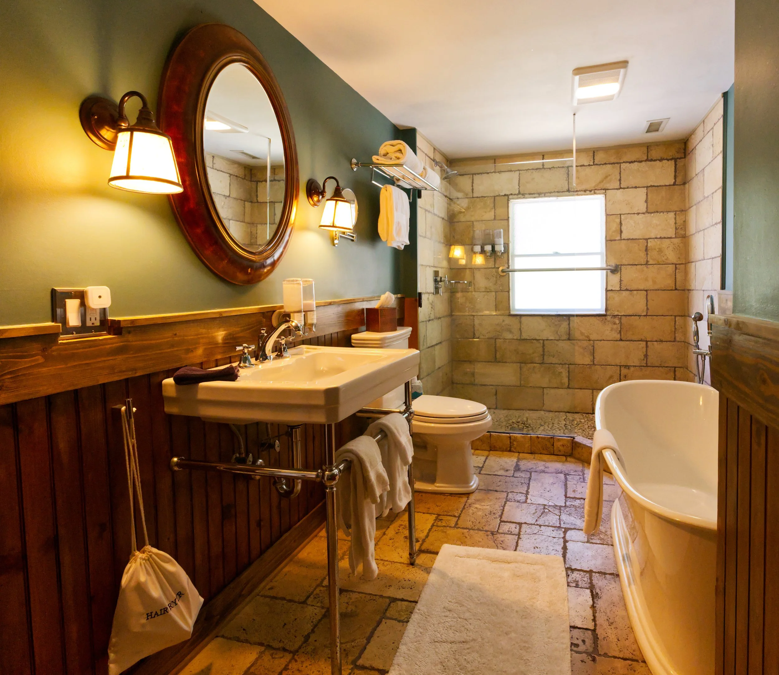 Bathroom with beige stone tiles, green wall, and wood vanity. Contains a white sink, toilet, bathtub, and various towels. Mirrors and wall-mounted lights above the sink. Window providing natural light.