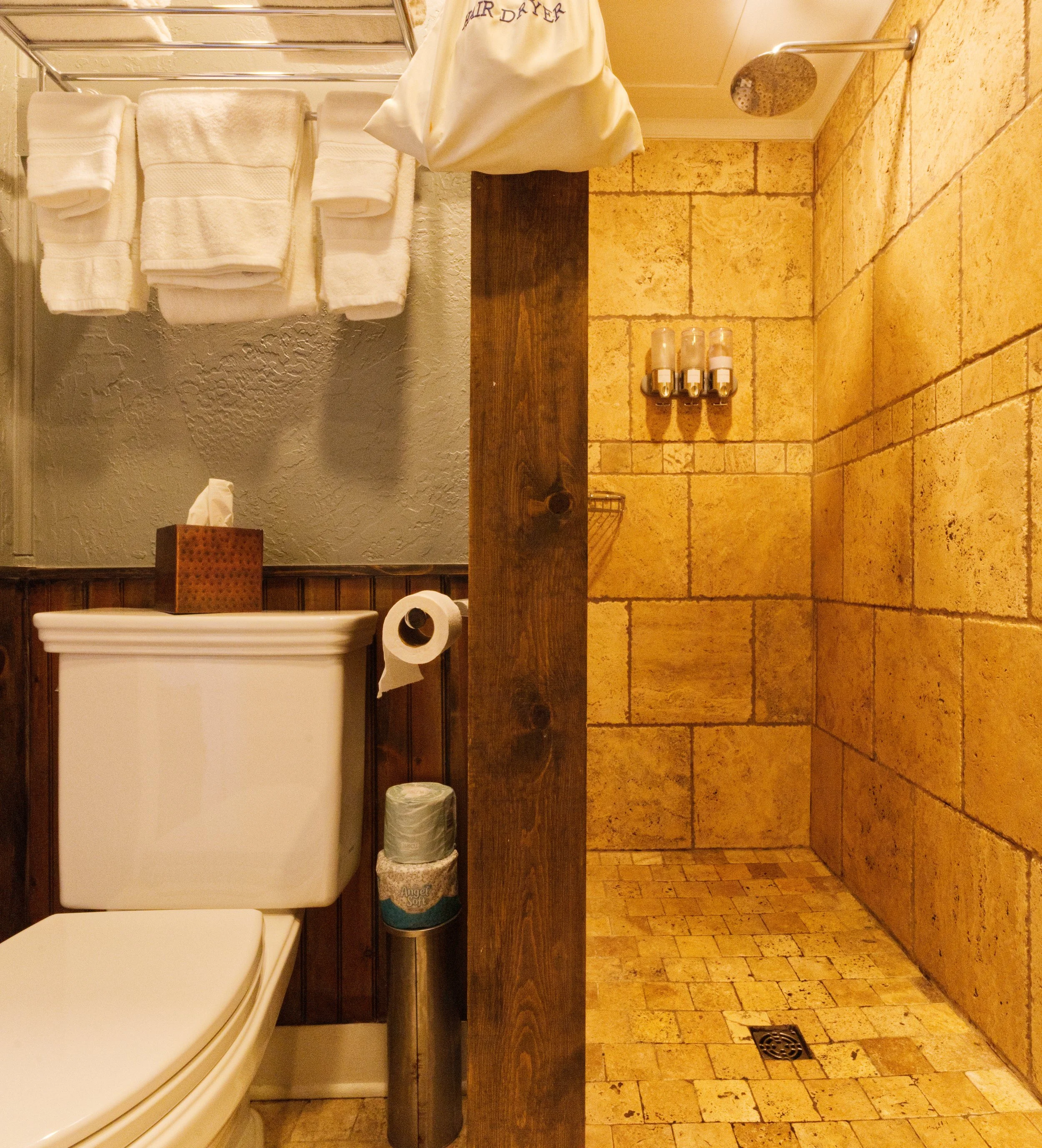A bathroom sink area with a white rectangular sink, a tissue box, a roll of toilet paper, and a trash can. Towels are neatly folded and stacked on a rack on the wall. There is a showerhead on the wall to the right and three soap dispensers mounted on the wall. The countertop has a wooden beam running across it. The floor and walls are made of beige tiles, and there is a drain in the floor near the corner.