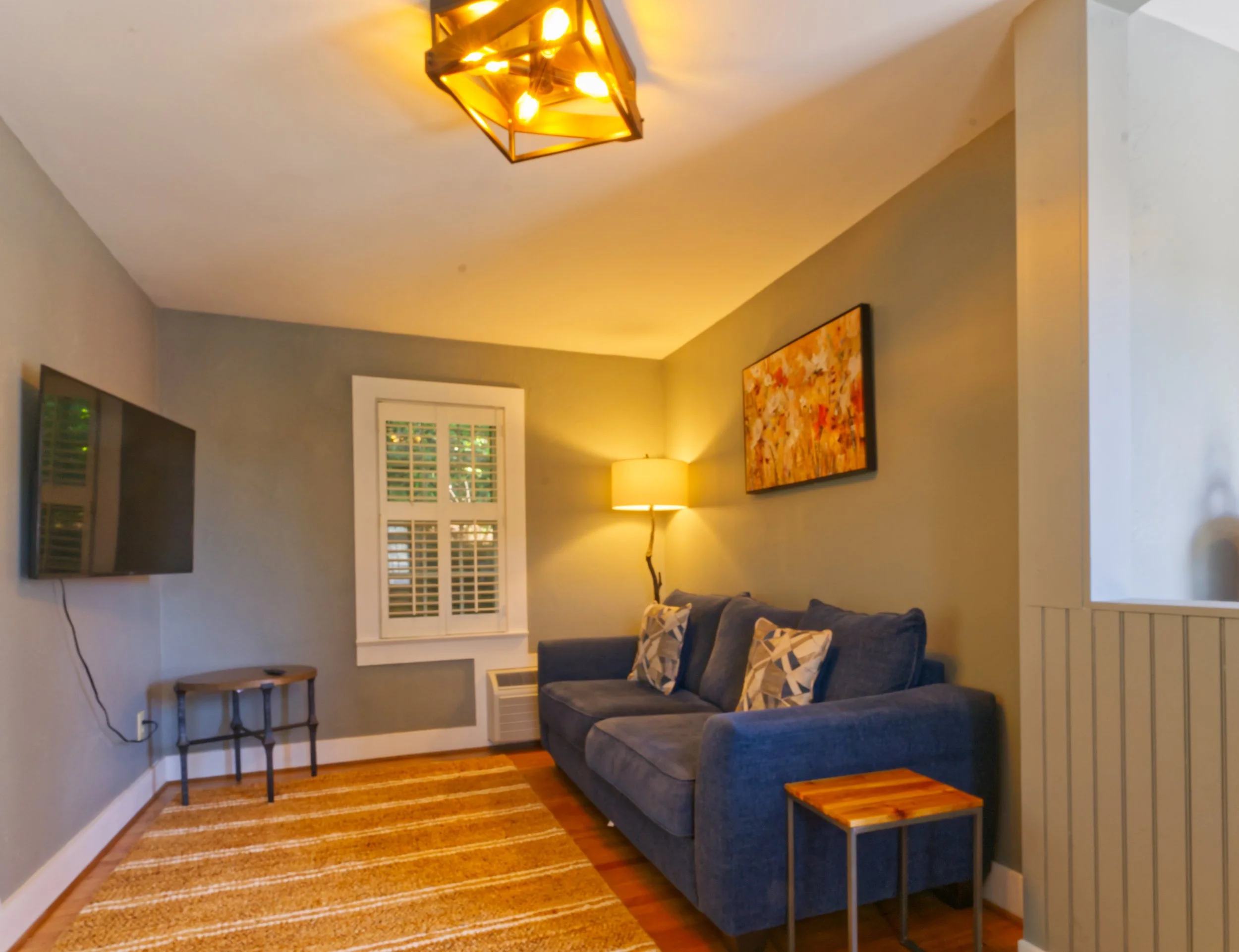Living room with a gray couch, colorful pillows, wooden side tables, a window with shutters, a wall-mounted TV, a colorful abstract painting, a floor lamp, and a modern ceiling light fixture.