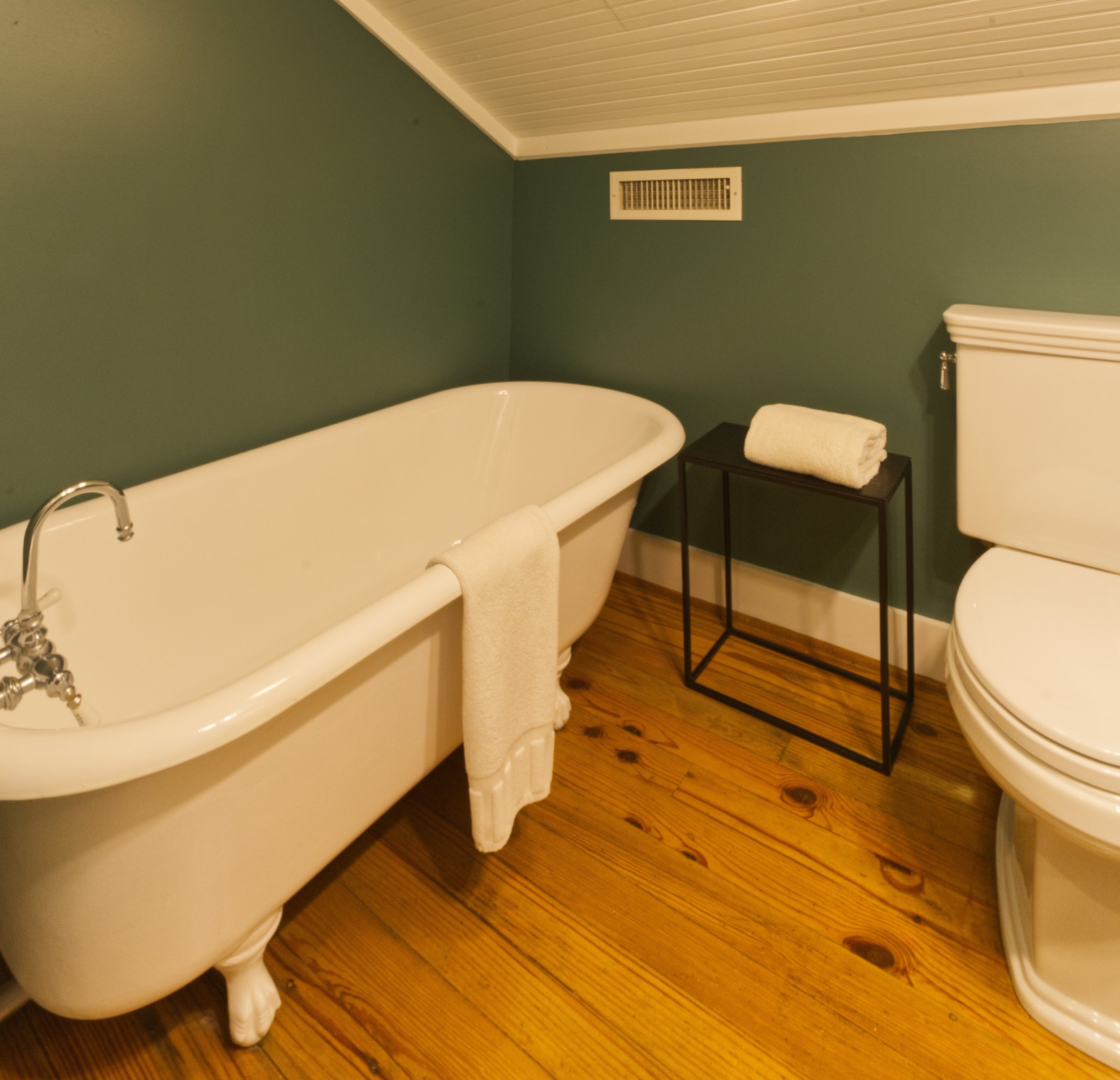 Bathroom with green walls, white bathtub, rolled towels on a small table, and a wooden floor.