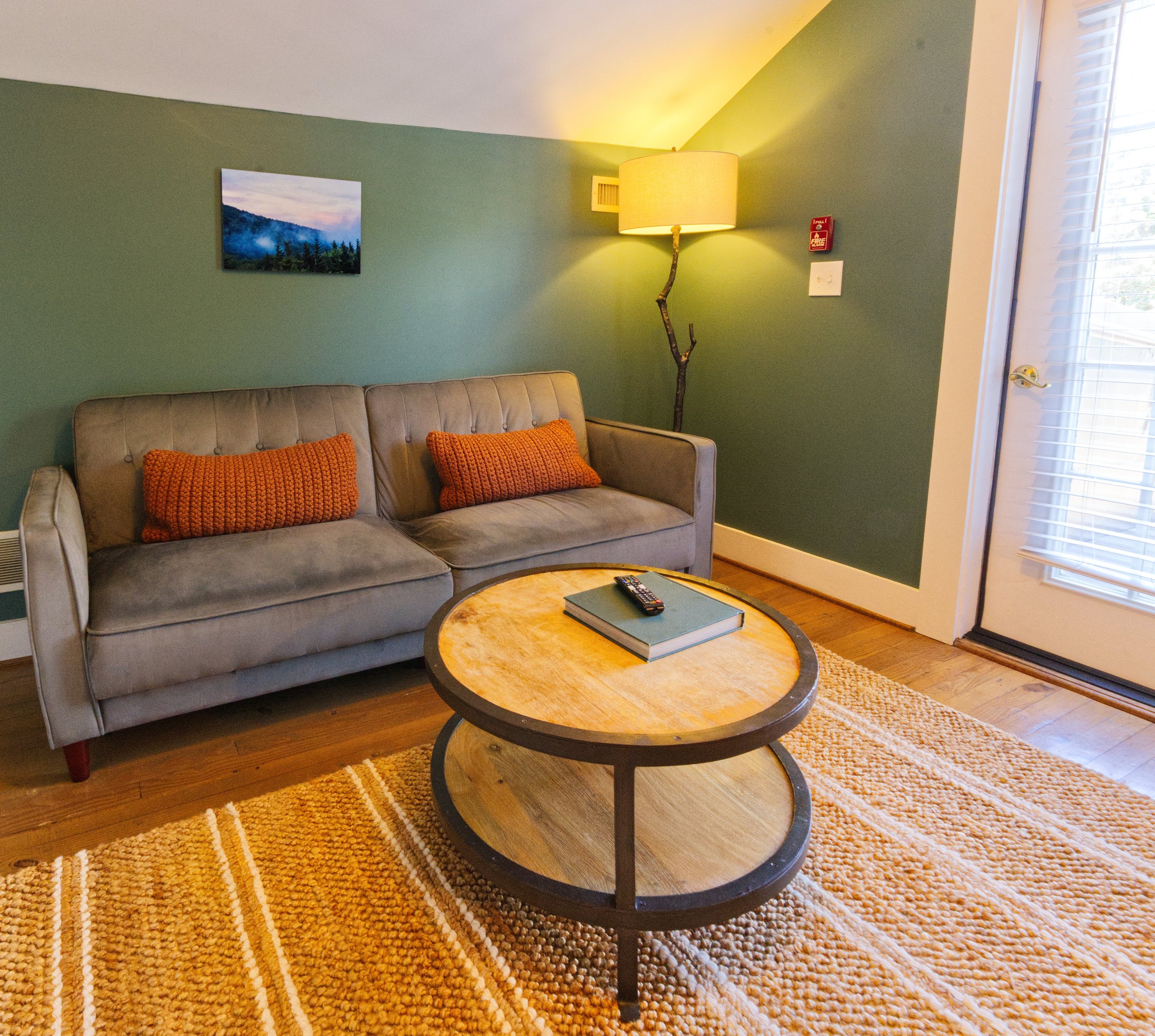 Living room with a green accent wall, beige couch with orange pillows, round wooden coffee table, floor lamp with a branch-style stand, a landscape photo on the wall, orange rug, and a window with blinds.