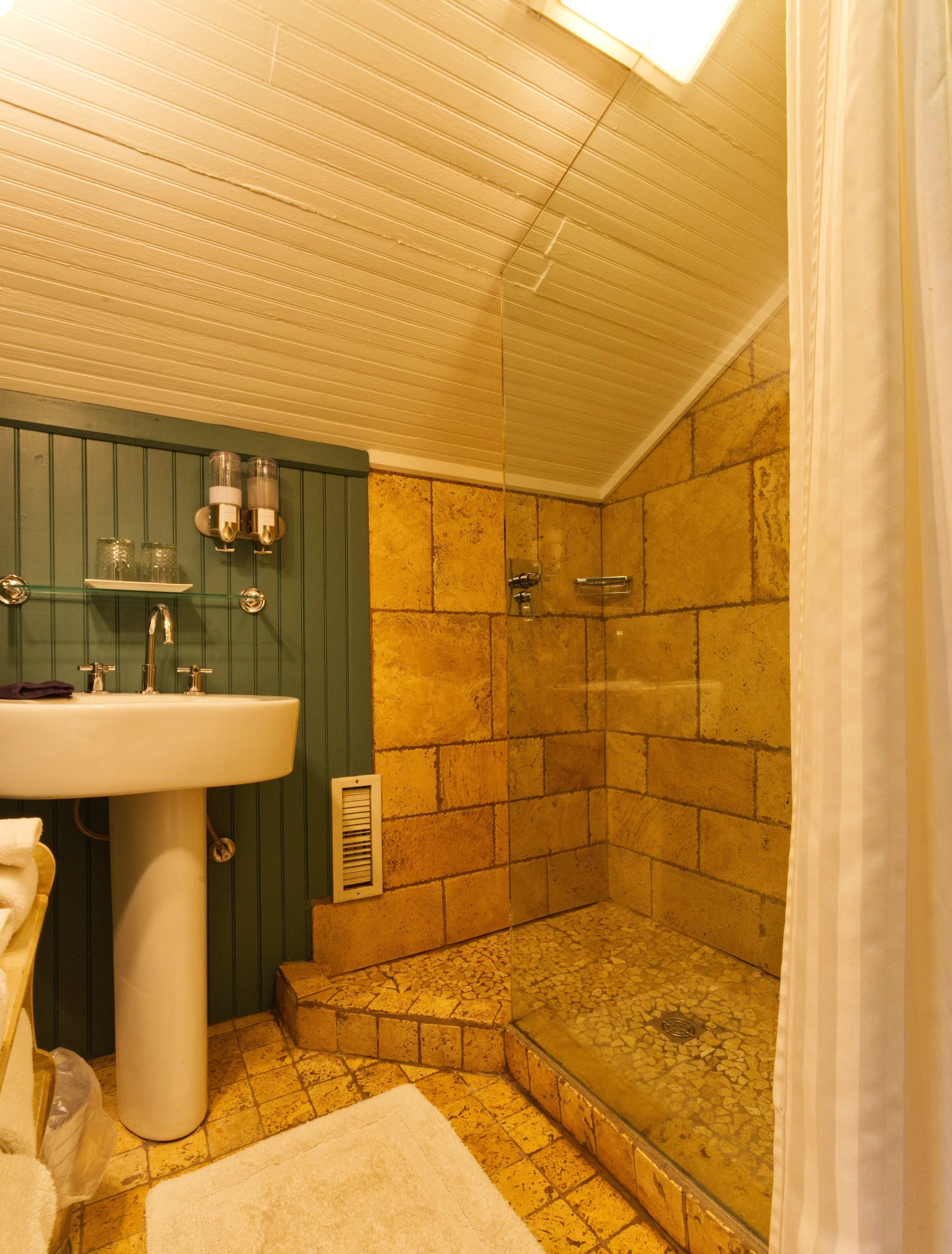 A bathroom with a pedestal sink, beige tile walls and floor, a shower area with a textured tiles floor and a curtain on the right side, and a green wooden cabinet with glass shelves and lotion dispensers on top.