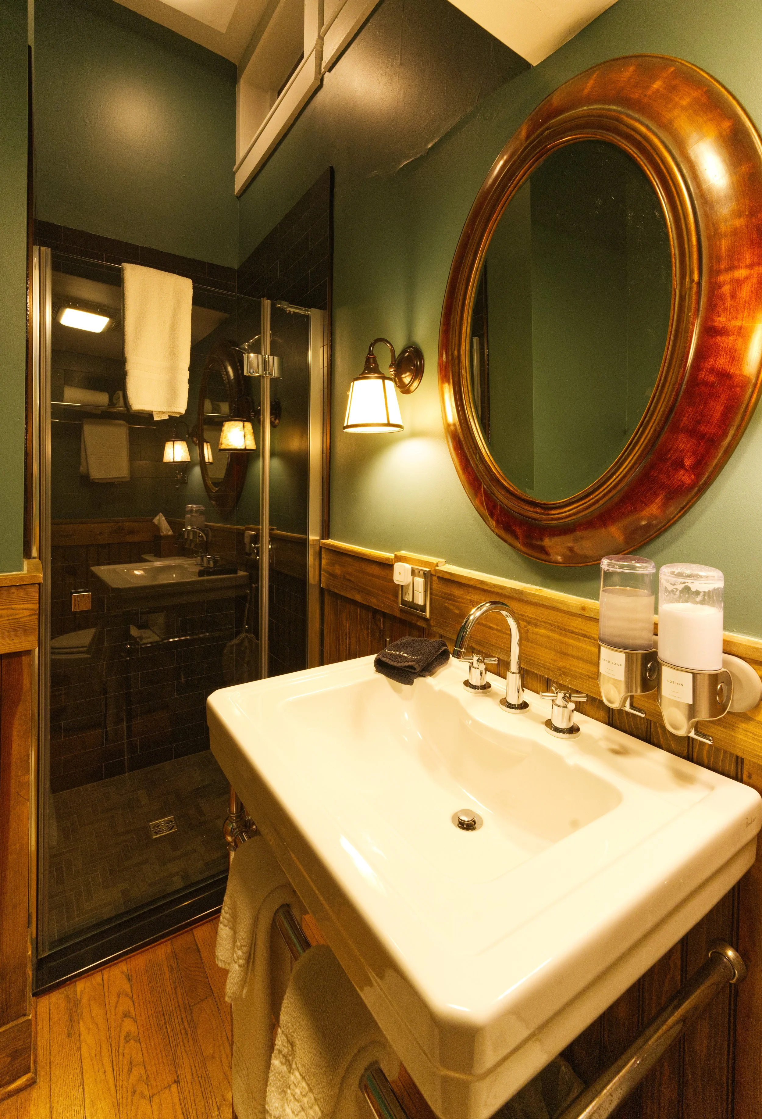 A bathroom with a vintage-style white sink with a towel hanging from it, mounted to a wooden wall beneath a large, oval, gilded mirror. To the right of the sink, there are wall-mounted soap and lotion dispensers. Behind the sink, a glass-enclosed shower has black tiles and a towel hung on the door. The wall is painted green, with a warm light fixture illuminating the space.