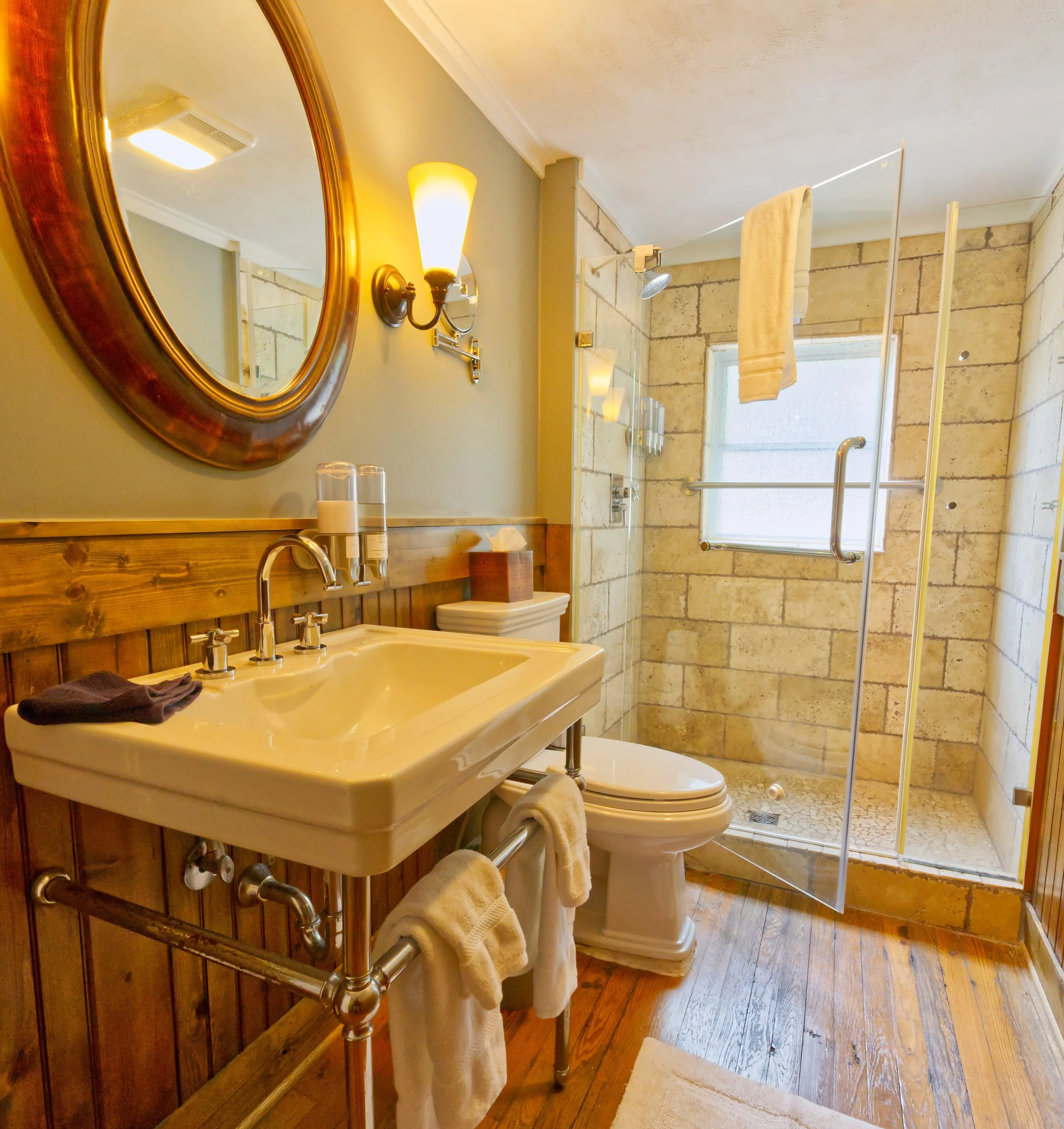 Bathroom with a wooden vanity, oval mirror with a wooden frame, wall lamp, beige tiles, glass shower door, towel hanging, and a window with a frosted glass. Wooden floor and a white rug.