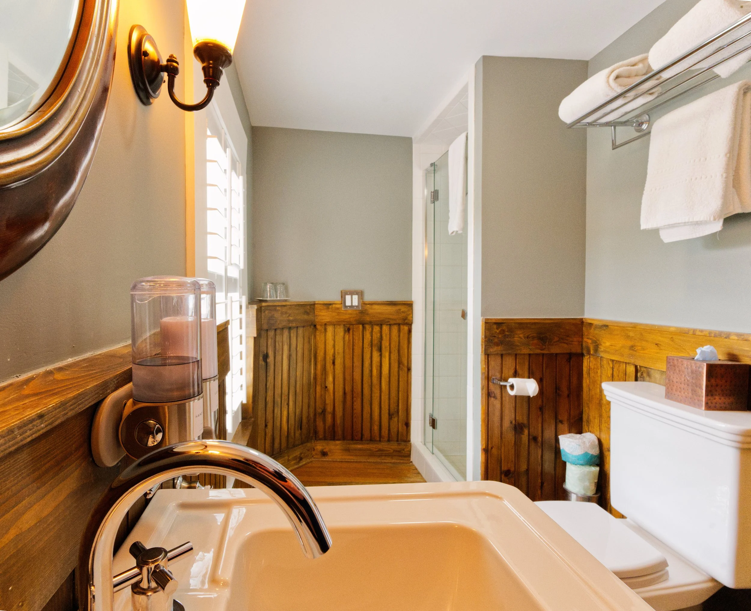 A bathroom with a wooden vanity, a white sink with a silver faucet, a wall-mounted black sconce light, a mirror, and towels on a metal towel rack. A view into a shower with a clear glass door and white tile is in the background. There are windows with white shutters and wooden trim.
