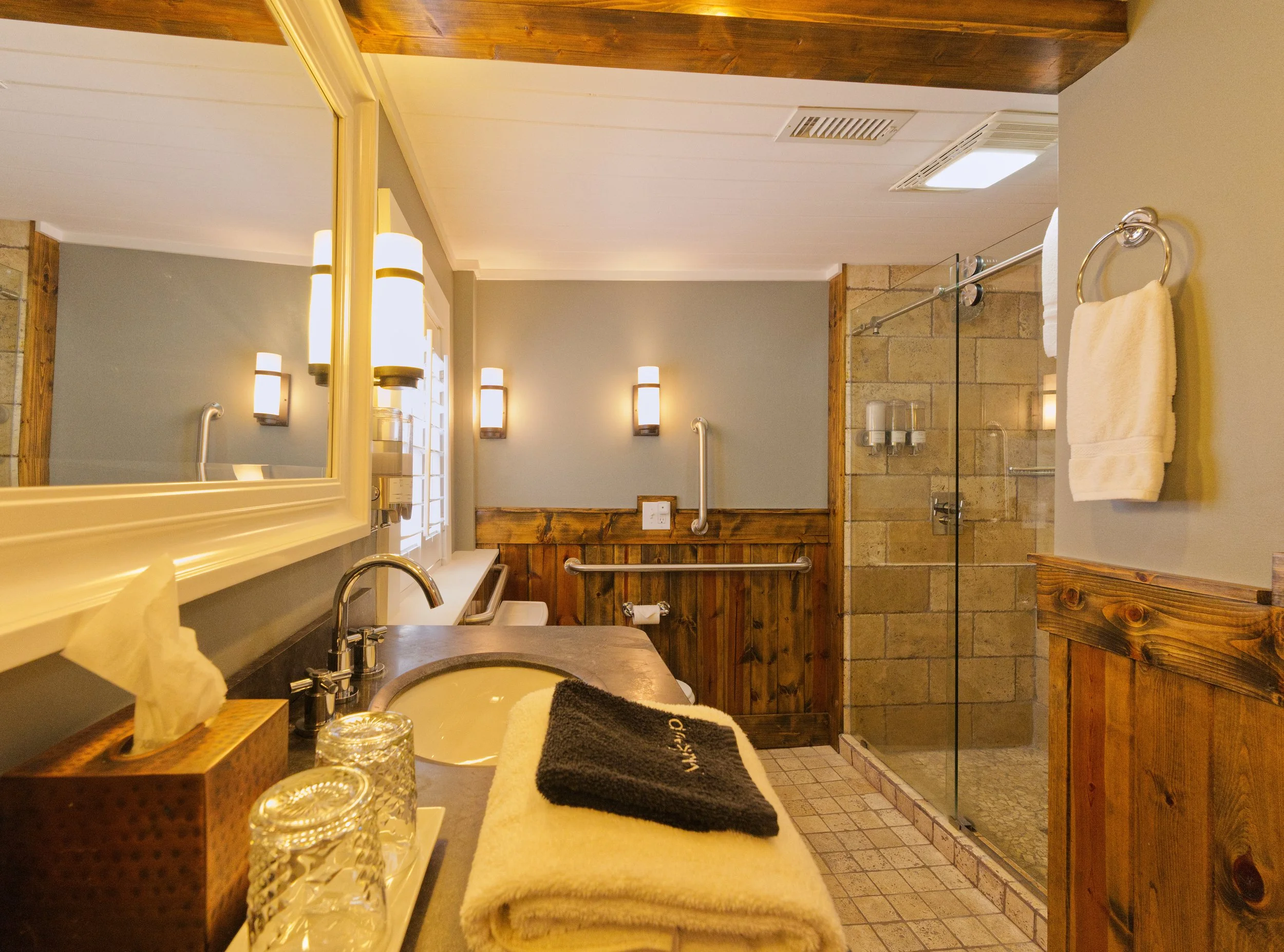 A bathroom with beige walls, wooden accents, and a glass-enclosed shower. There are four wall-mounted lights above a mirror, a white countertop with a sink, a black towel, and decorative glass containers. A handrail and a white towel are visible, and the ceiling has a light fixture and vent.