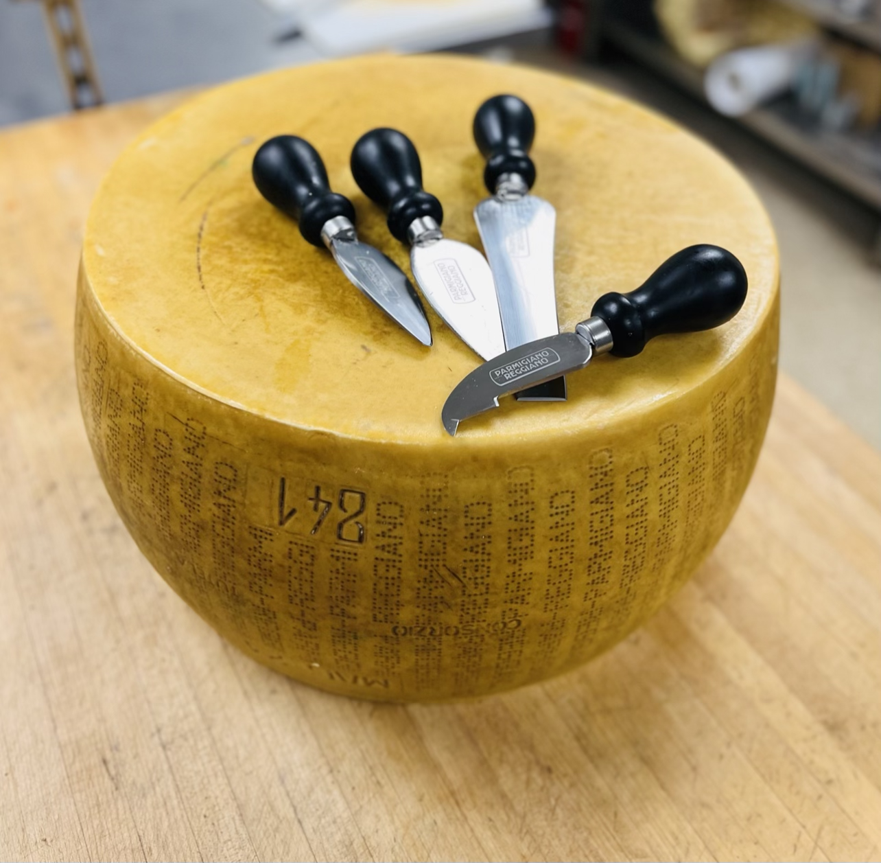 A large wheel of Parmigiano-Reggiano cheese with four cheese knives resting on top, on a wooden surface in a cheese shop or kitchen.