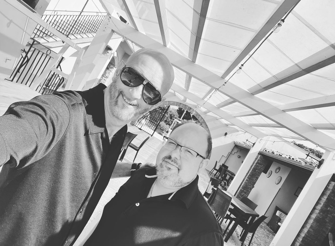 Two men taking a selfie outdoors on a patio area, with chairs and tables, under a pergola with string lights, in black and white.