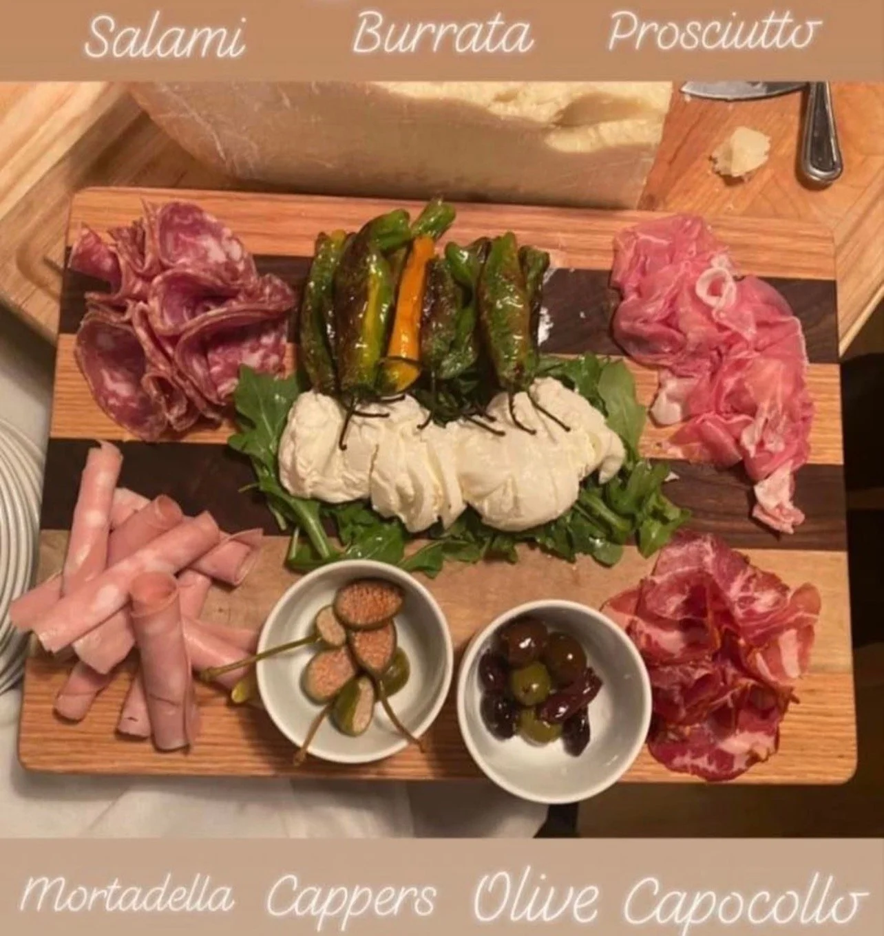 Wooden platter with an assortment of Italian cured meats, cheeses, roasted peppers, and small bowls of olives and pickles, labeled with the varieties: salami, burrata, prosciutto; also includes mortadella, capers, and olive capocollo.
