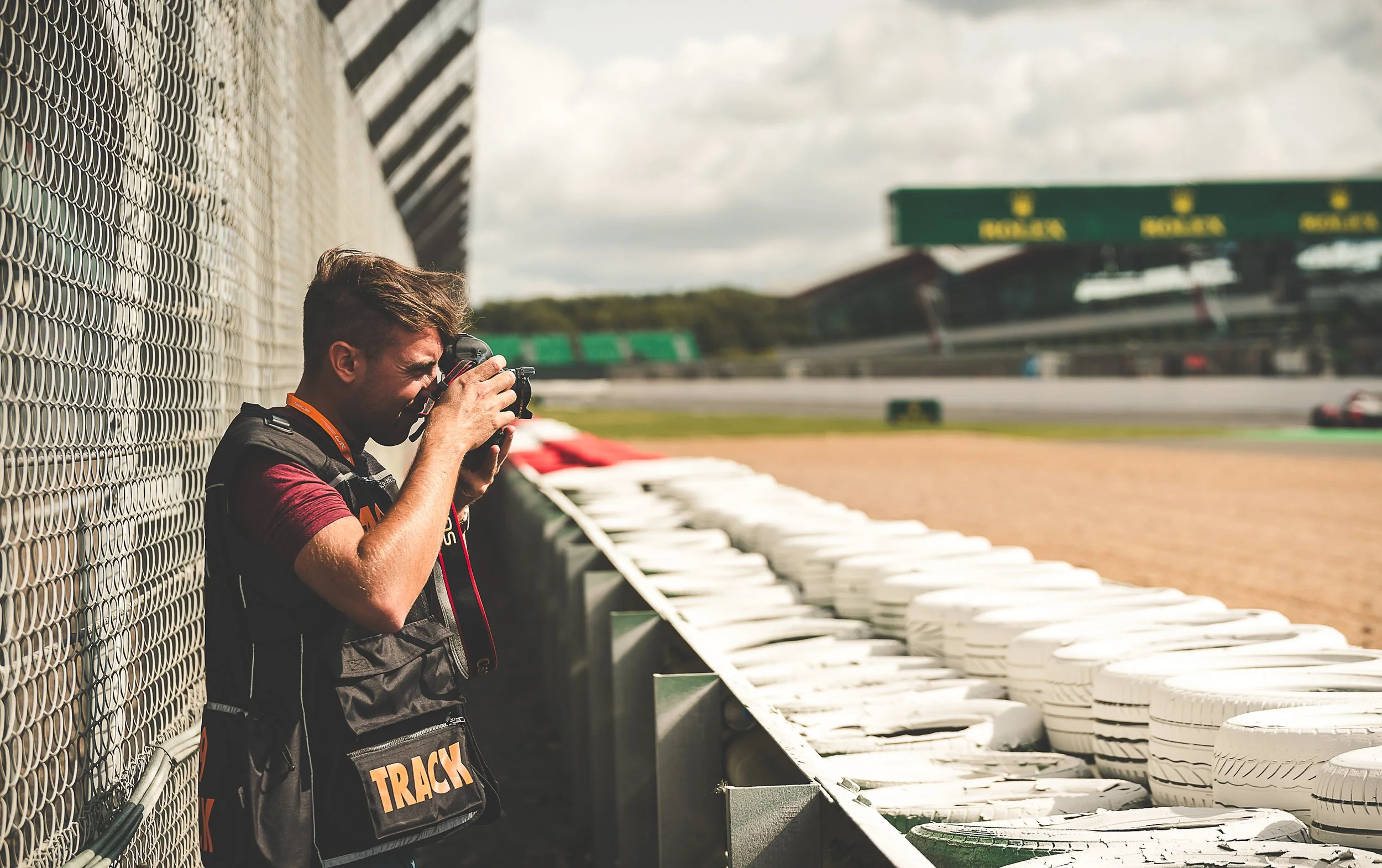 Photographer Mark J Timms taking pictures at a racetrack, standing beside a chain-link fence with tires stacked along the barrier, and race cars on the track in the background.