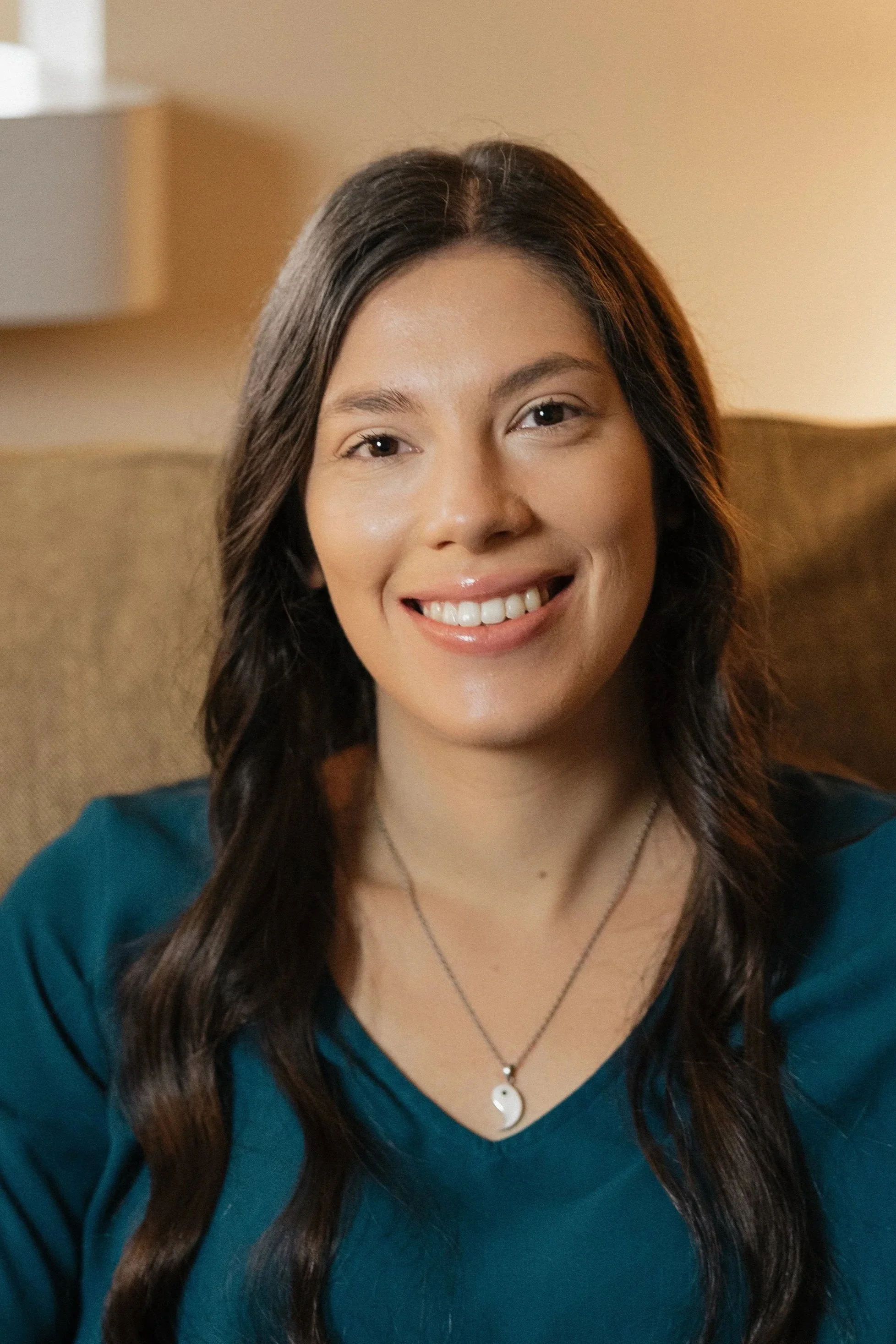 A woman with long dark hair smiling at the camera, wearing a teal top and a yin-yang necklace, sitting on a beige sofa in a cozy indoor setting.