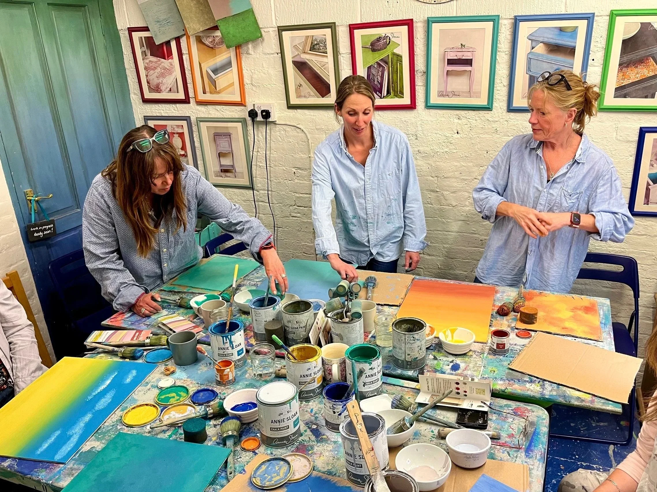 Three women are standing around a table covered with cans and bowls of paint, with several colorful framed pictures of painted furniture on the wall behind them, in an art studio or workshop setting.