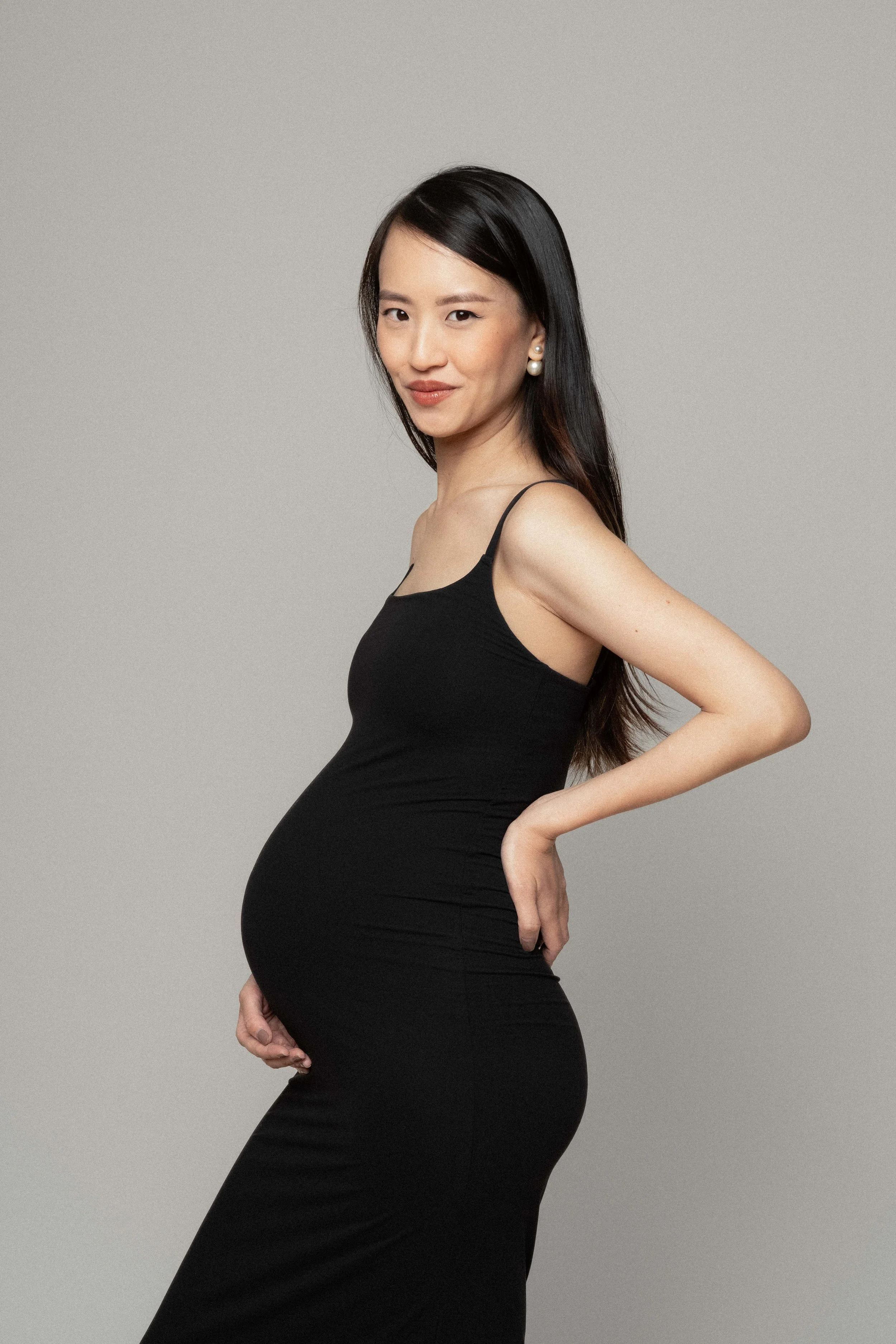 Christina in a black dress, maternity portrait against a neutral backdrop