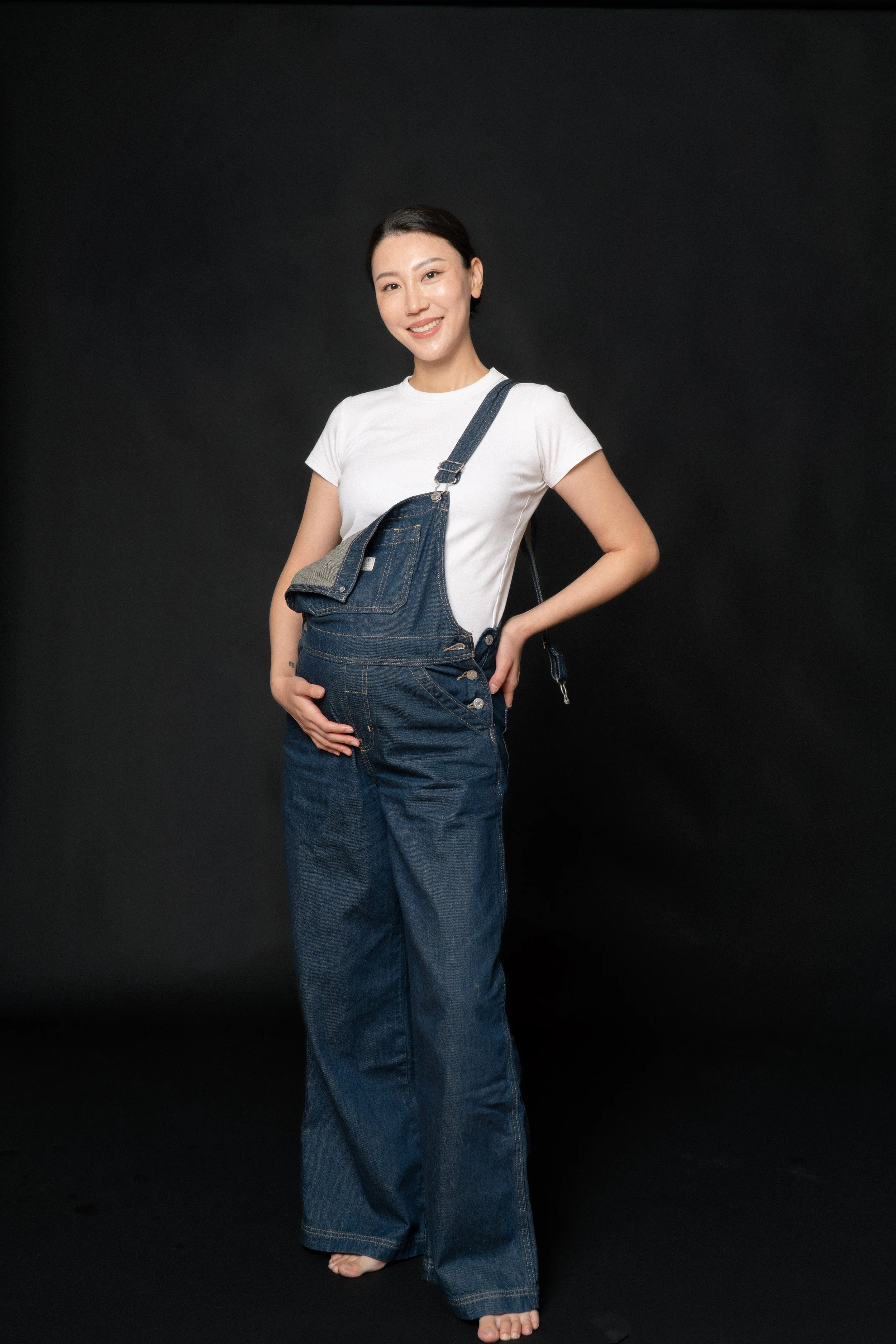 Cloris cradling her baby bump in soft studio light in a denim overall, full length