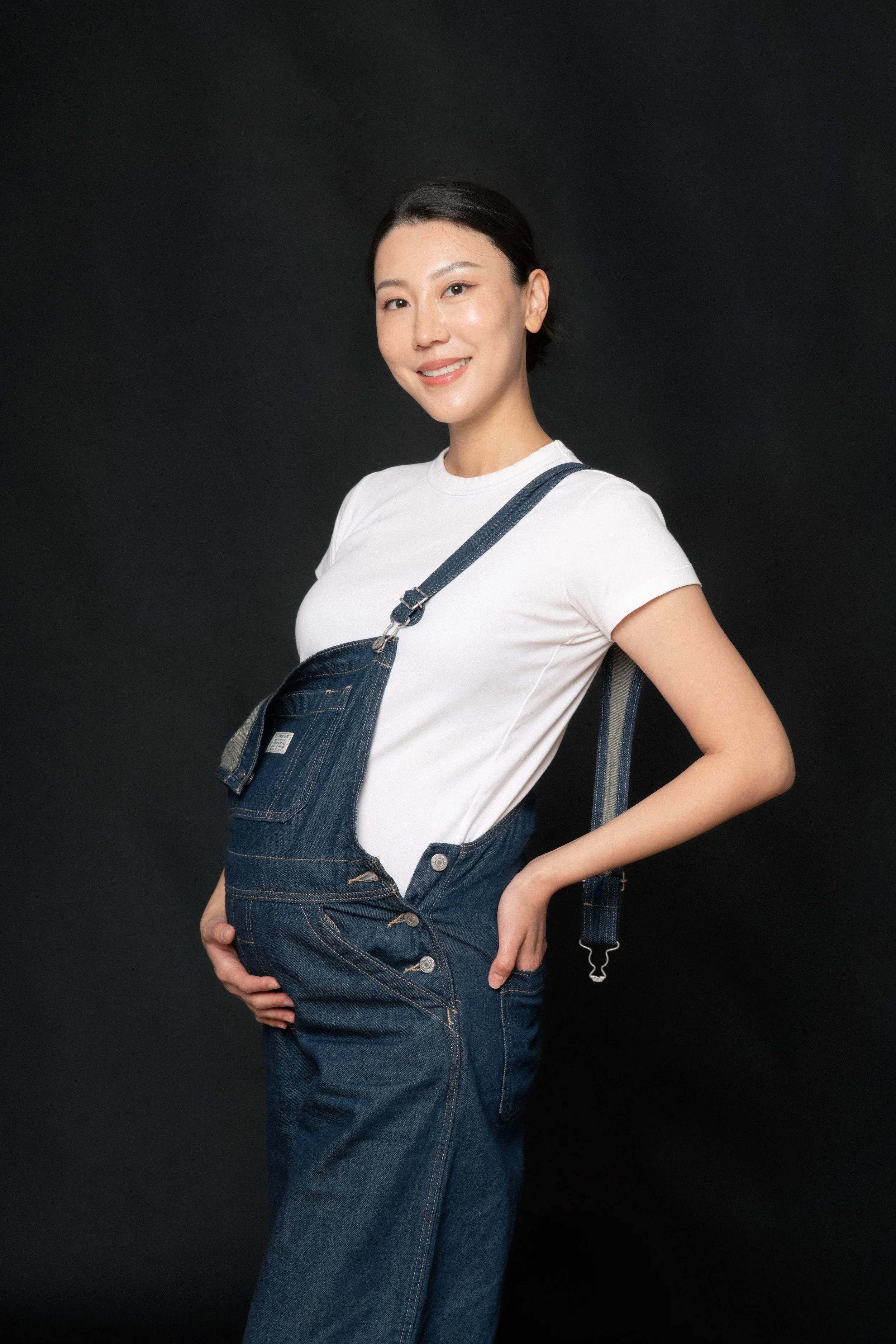 Cloris posing with her baby bump in soft studio light in a denim overall