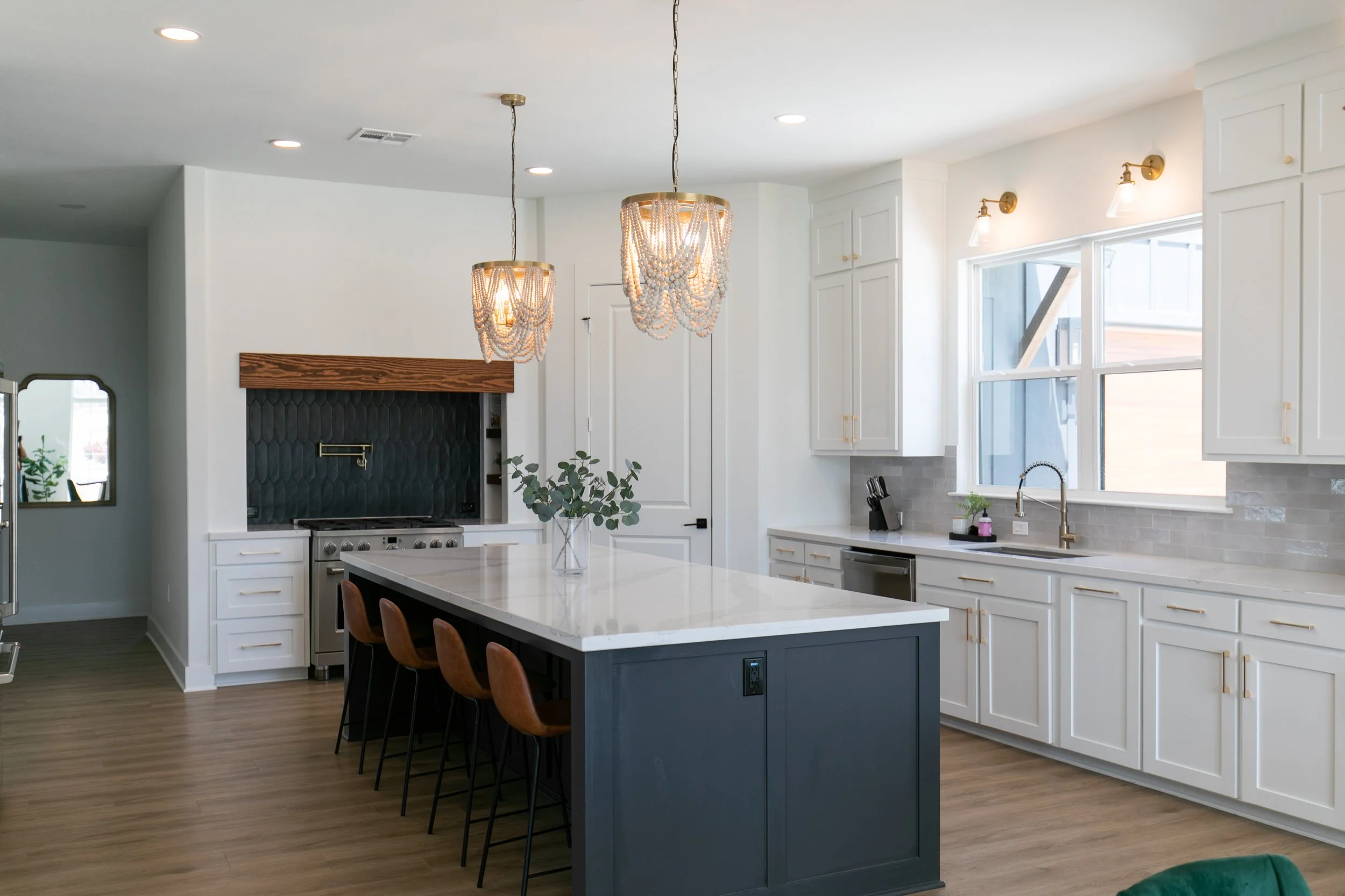 Modern kitchen with white cabinets, a large island with a dark base, pendant lighting, and a chandelier. There is a window above the sink, and a fireplace with a wooden mantle.