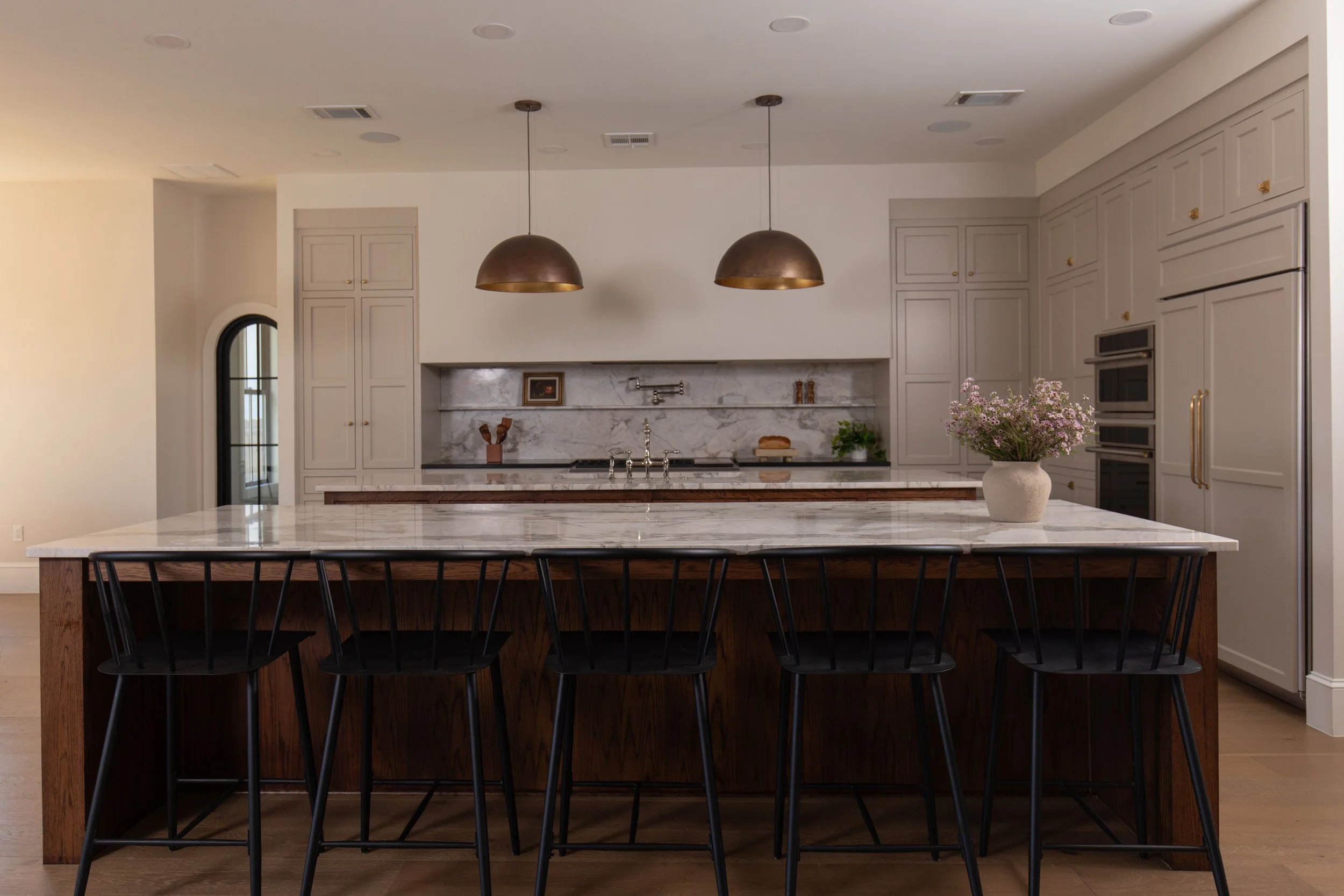 Modern kitchen with white cabinets, marble backsplash, and a large island with a wooden base and marble countertop. Hanging pendant lights above the island, a potted plant, and a vase with pink flowers on the counter.