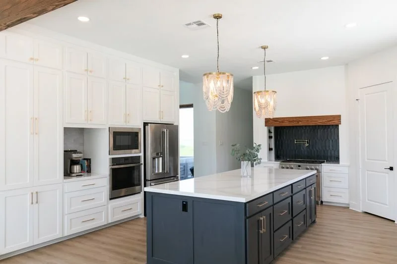 Modern kitchen with white cabinets, a navy blue kitchen island, and a stainless steel refrigerator. Features pendant lights above the island and a built-in oven and microwave. Decor includes a potted plant and a coffee maker.