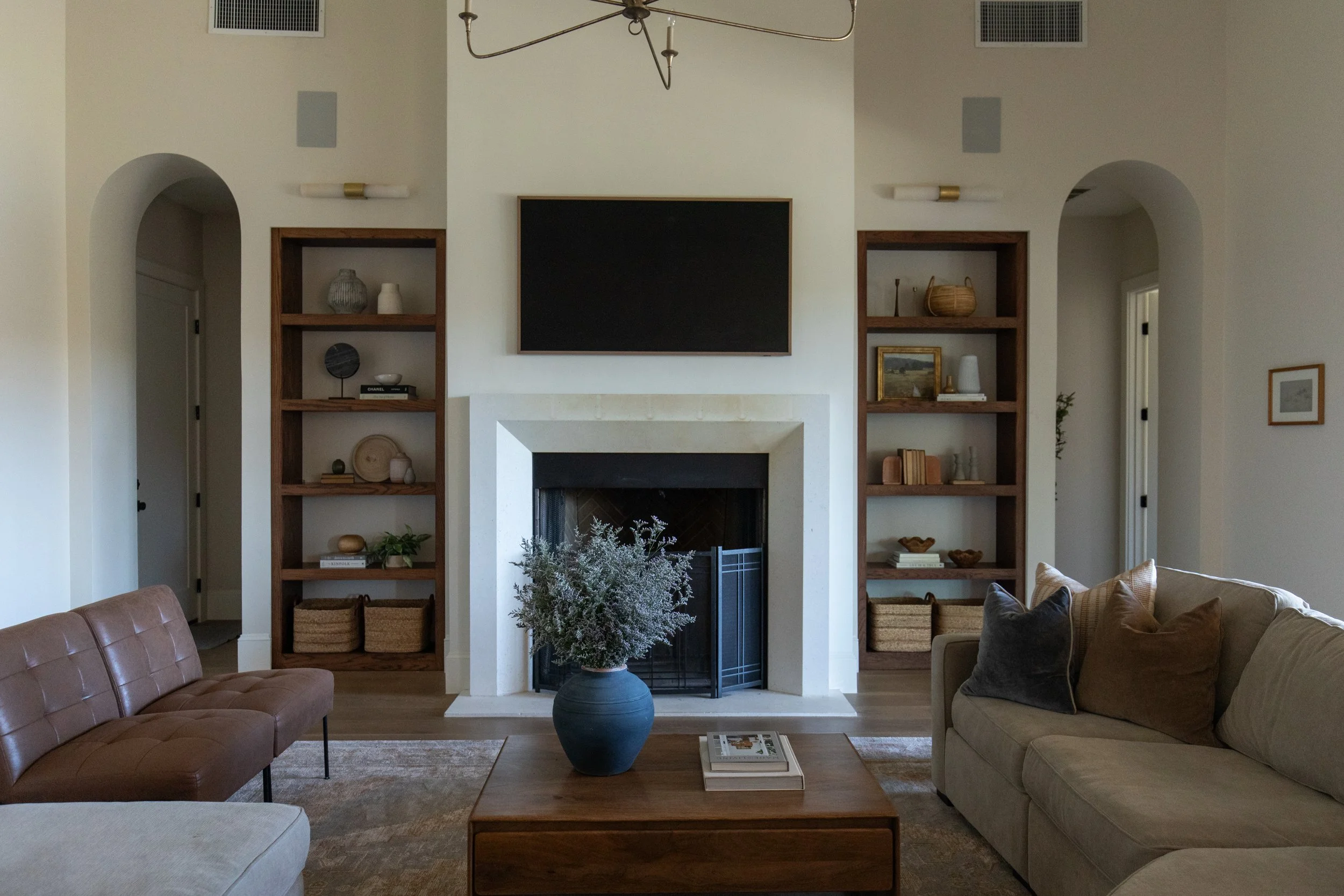 A living room with a white fireplace, two wooden bookshelves with decor, a black television above the fireplace, a beige sofa, a brown leather sofa, a coffee table with magazines, and a large patterned rug. There are decorative pillows on the sofas and a plant in a large gray pot near the fireplace.