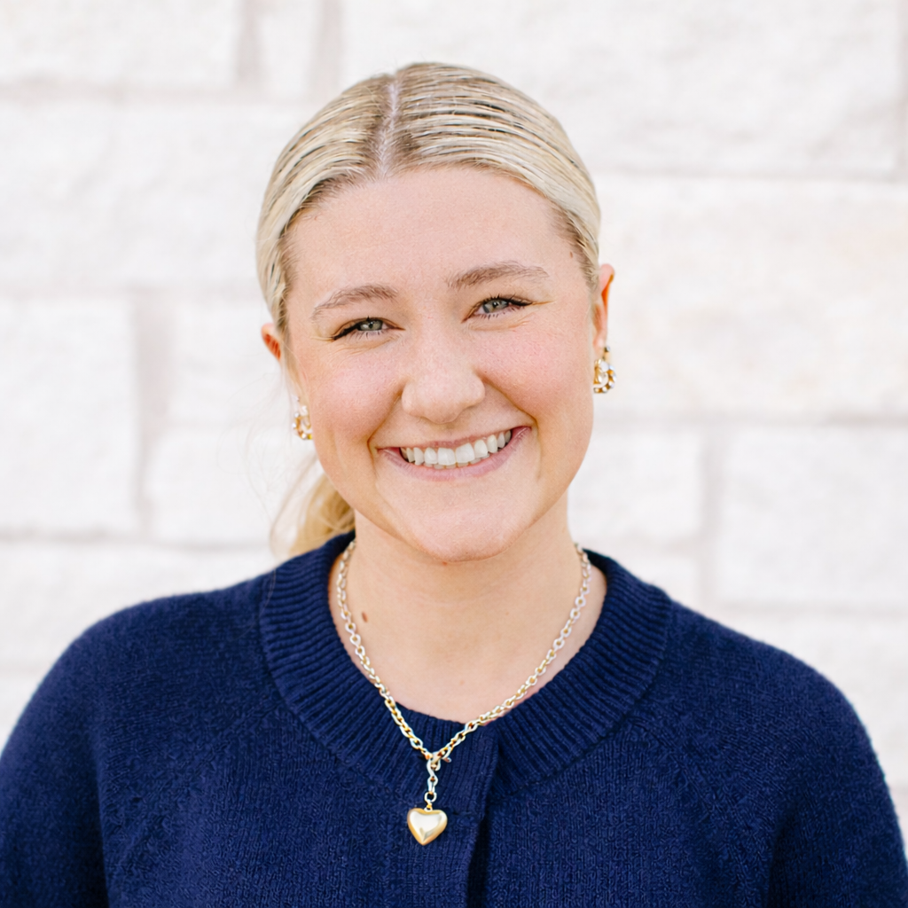 Portrait of a smiling woman with blonde hair tied back, wearing pearl earrings, a gold heart necklace, and a navy blue sweater with a white brick wall background.