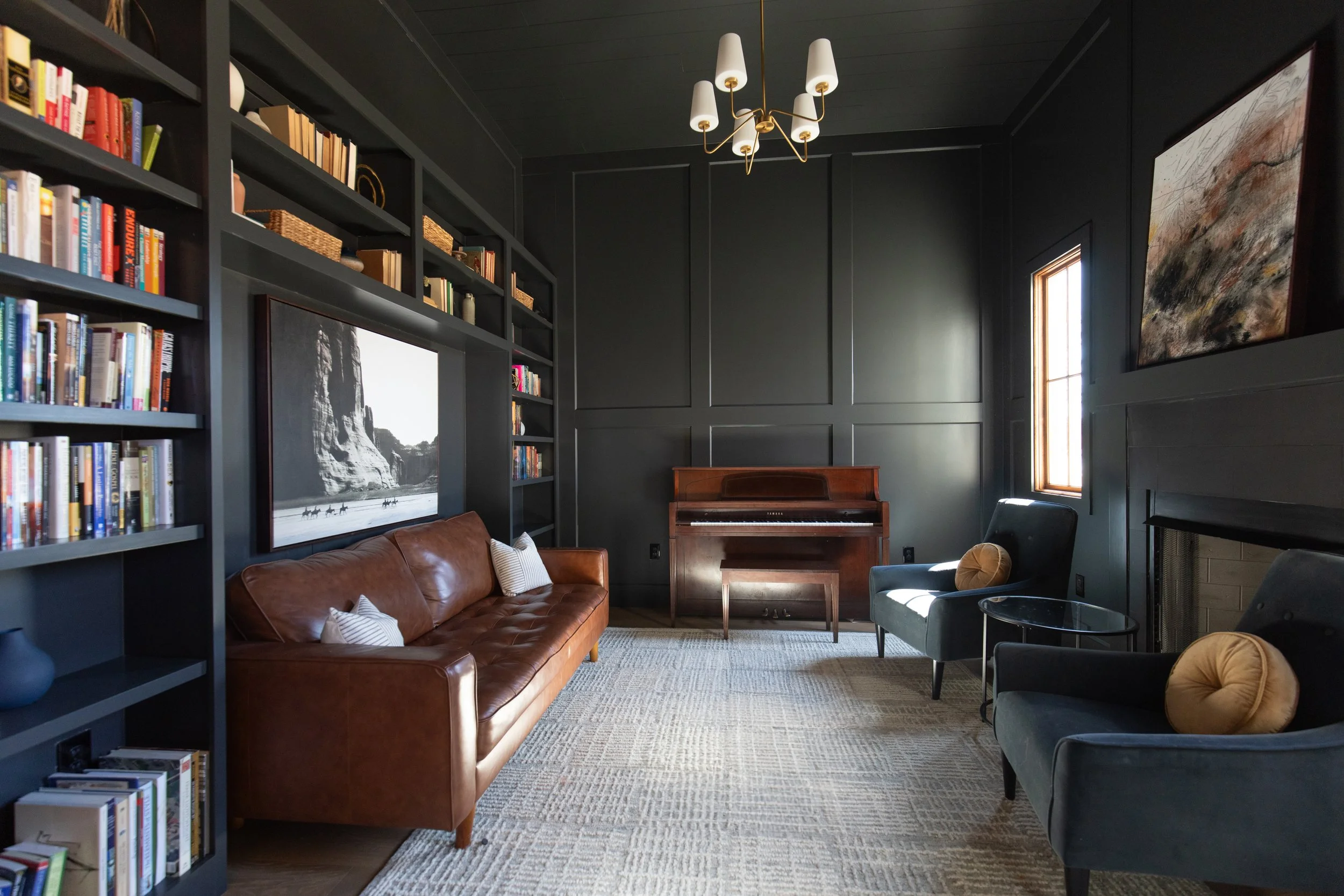 A living room with dark gray walls and ceiling. It features a brown leather sofa with pillows, a bookshelf filled with books, a charcoal armchair, a standing guitar, a framed black-and-white landscape photograph on the wall, and a large abstract painting. Small tables are placed near the chairs, and a window lets in natural light. A modern chandelier hangs from the ceiling.