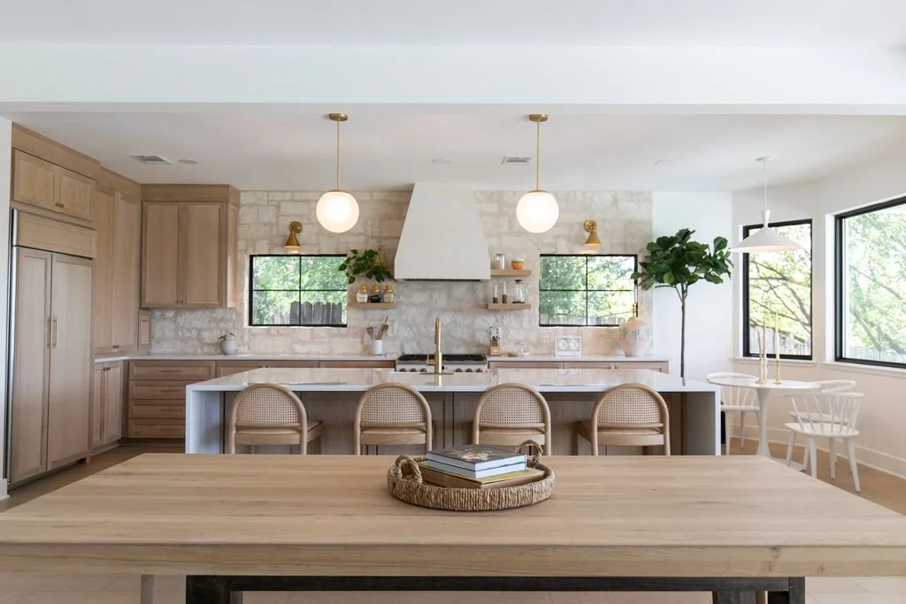 Bright, modern kitchen with light wood cabinetry, a large island with seating, white stone backsplash, two round hanging lights, large windows with greenery outside, and a dining area with white chairs and a small round table.