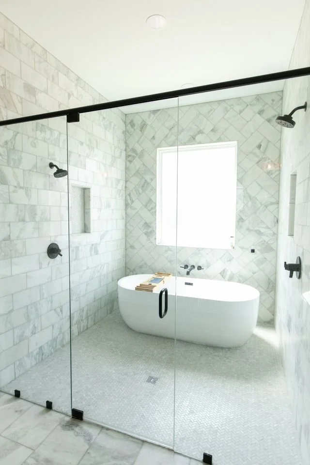 Modern bathroom with marble walls, glass shower enclosure, freestanding bathtub, and large window.