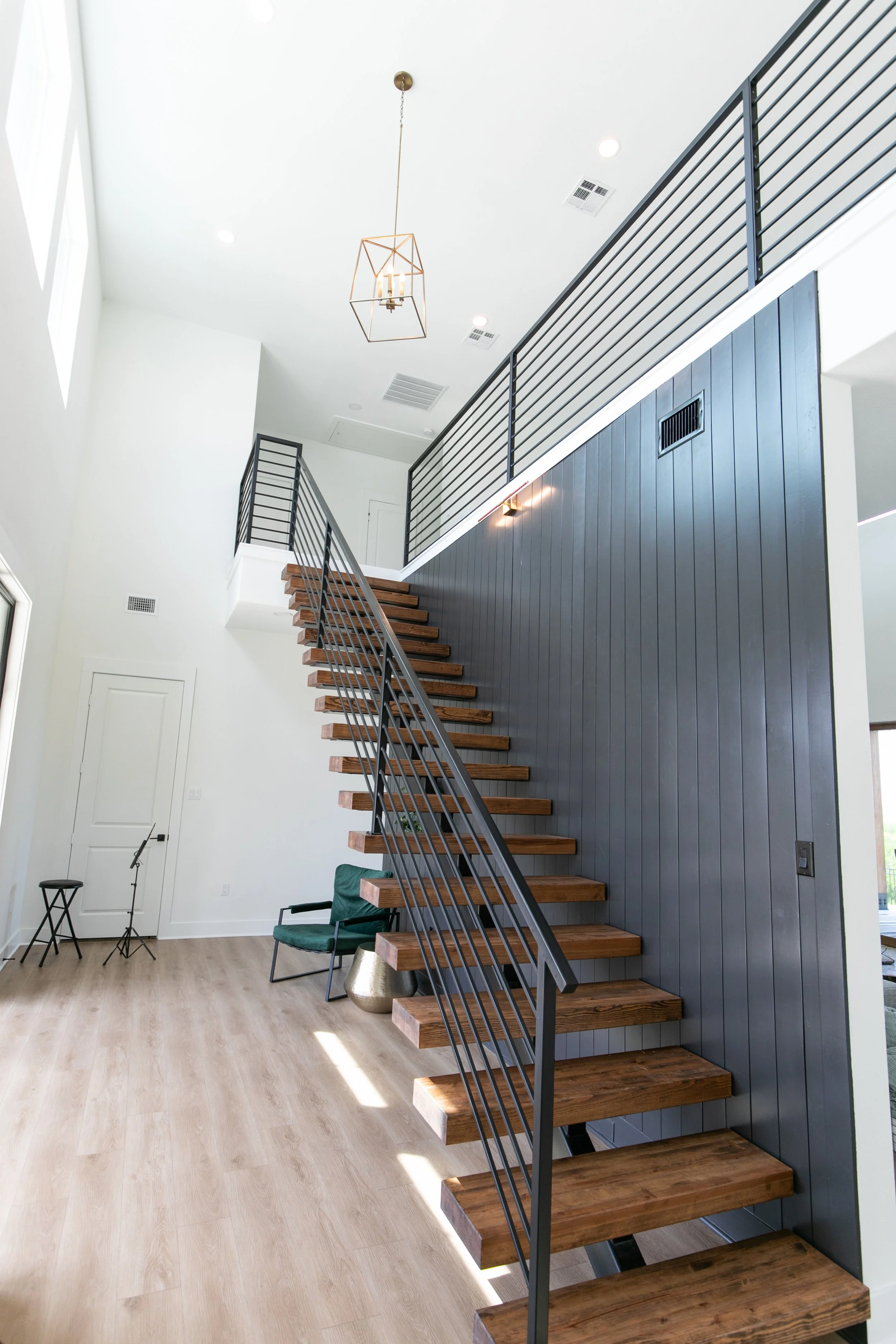 Interior view of a modern house featuring a staircase with wooden steps and a metal railing, a blue accent wall, a green armchair, a small stool, a metallic vase, a black chair, and a wall-mounted light fixture. There is a large window on the left, and the ceiling has recessed lighting and a geometric pendant light.