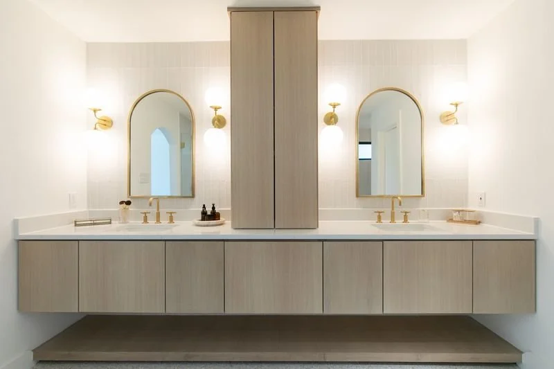 Double vanity bathroom with two arched mirrors, gold fixtures, light wall sconces, and a central storage cabinet.