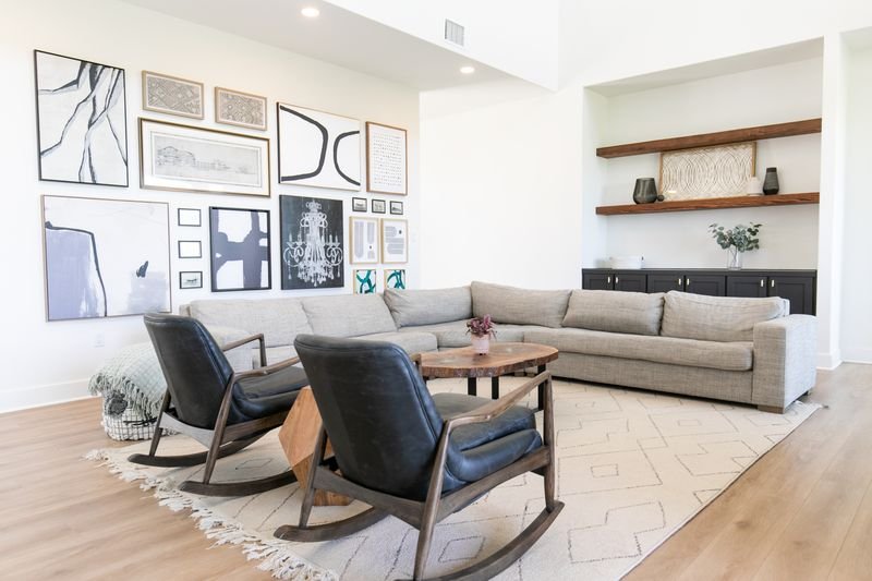 Living room with beige sectional sofa, two black leather armchairs around a wooden coffee table, wall art collage, wooden wall shelves with decorative items, light-colored area rug, hardwood flooring.