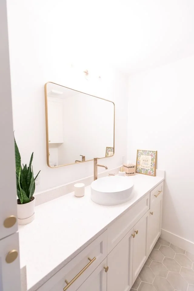 Bathroom vanity with white countertop, oval vessel sink, rectangular mirror, gold fixtures, potted snake plant, framed decorative art, soap dispenser, and candle, with light-colored tiled floor.