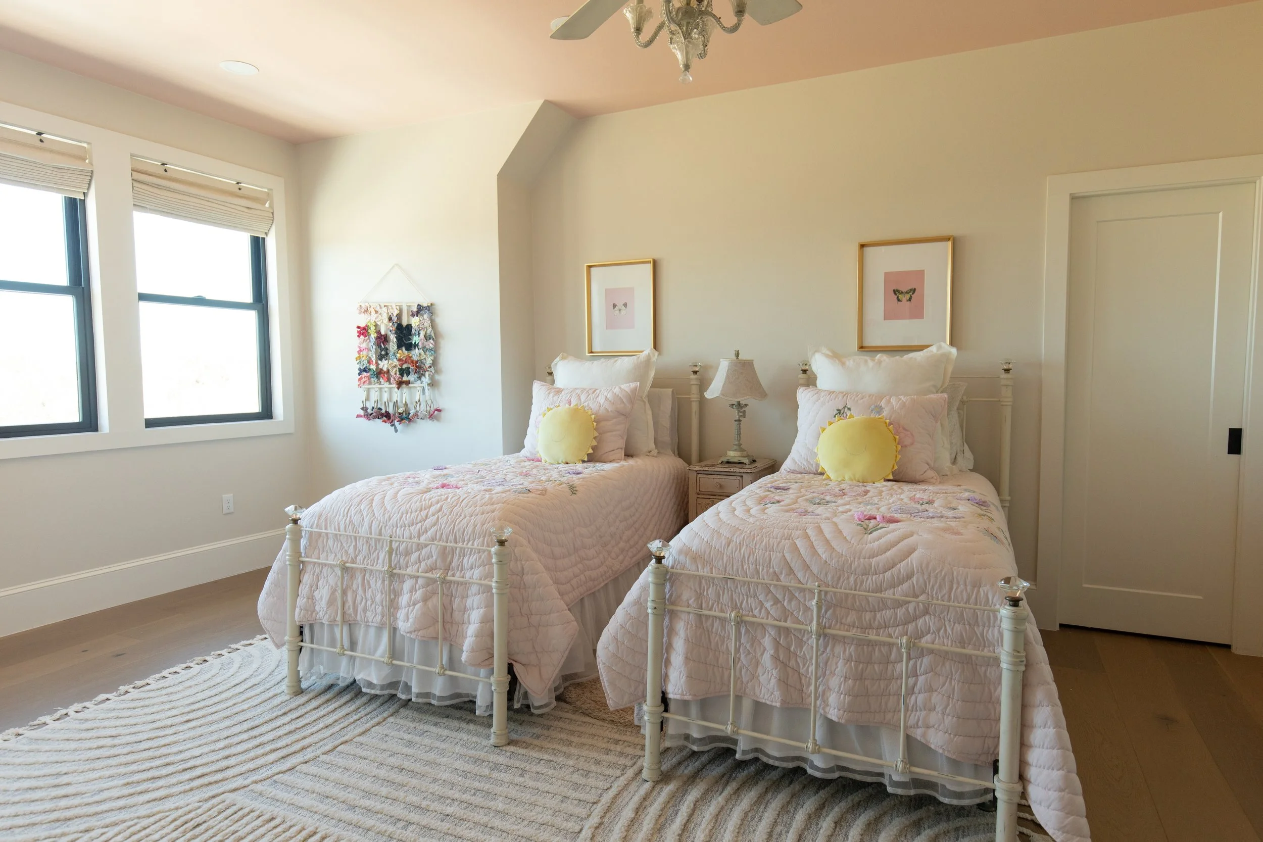 A bedroom with two twin beds with white metal frames and pink quilted bedspreads with floral patterns. Each bed has a round yellow pillow. There are two framed butterfly pictures above the beds and a nightstand with a vintage lamp between them. The room has three windows with white blinds, a woven wall hanging, and a white ceiling with a chandelier.
