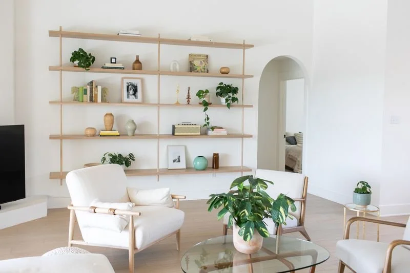 Living room with white walls, wooden flooring, and minimalist decor. There are white armchairs, a glass coffee table with a houseplant, and built-in wooden shelves with various decorative items. An archway leads to another room.