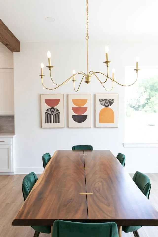Modern dining room with a large wooden table, green chairs, a brass chandelier, and abstract art prints on white walls.
