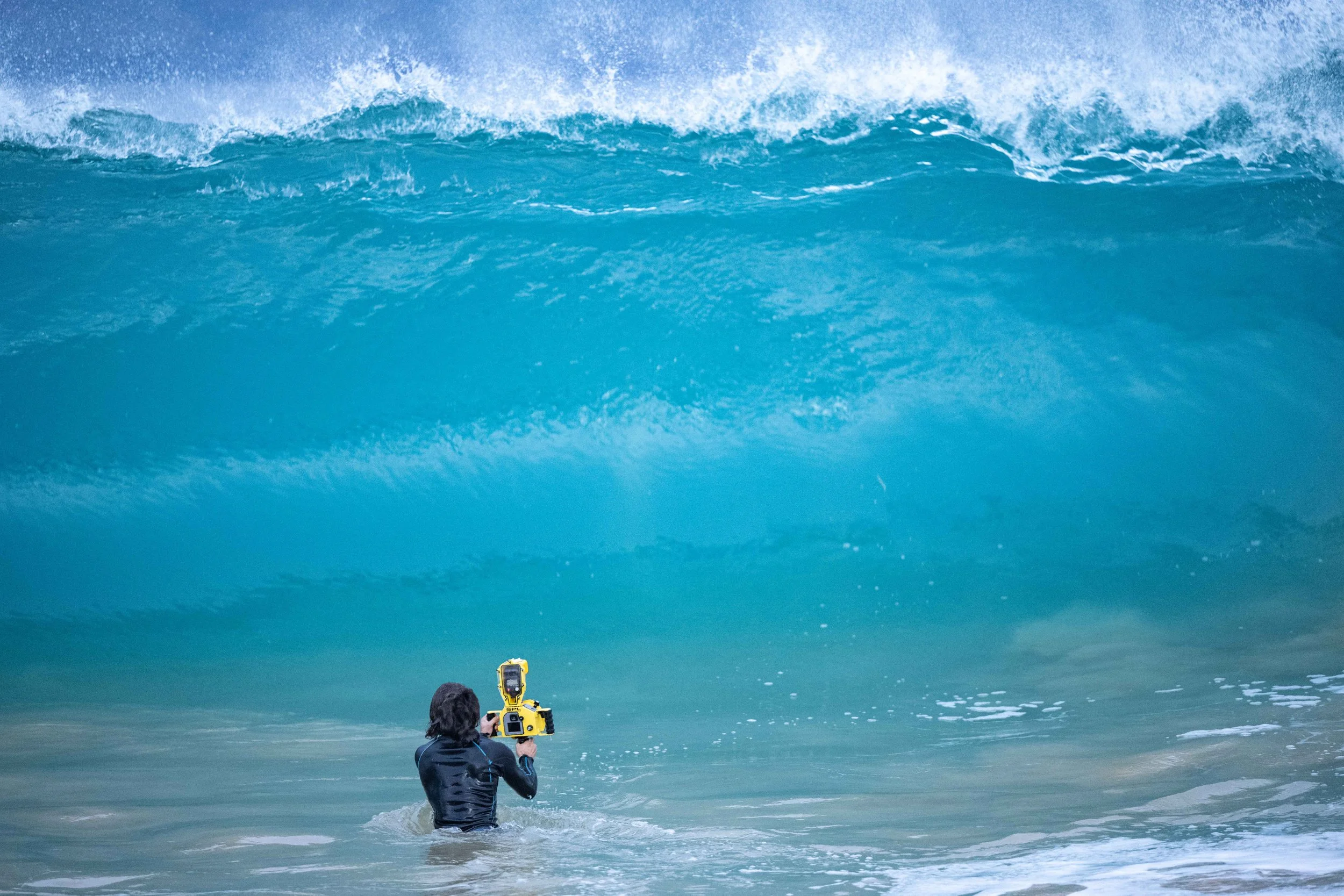 A person in black wetsuit holding a yellow camera underwater near a large blue wave