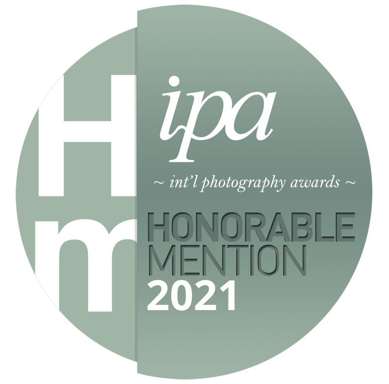 A circular badge design for the 2021 IPA International Photography Awards, featuring the text 'Honorable Mention 2021' in a modern, minimalist style with shades of green and gray.