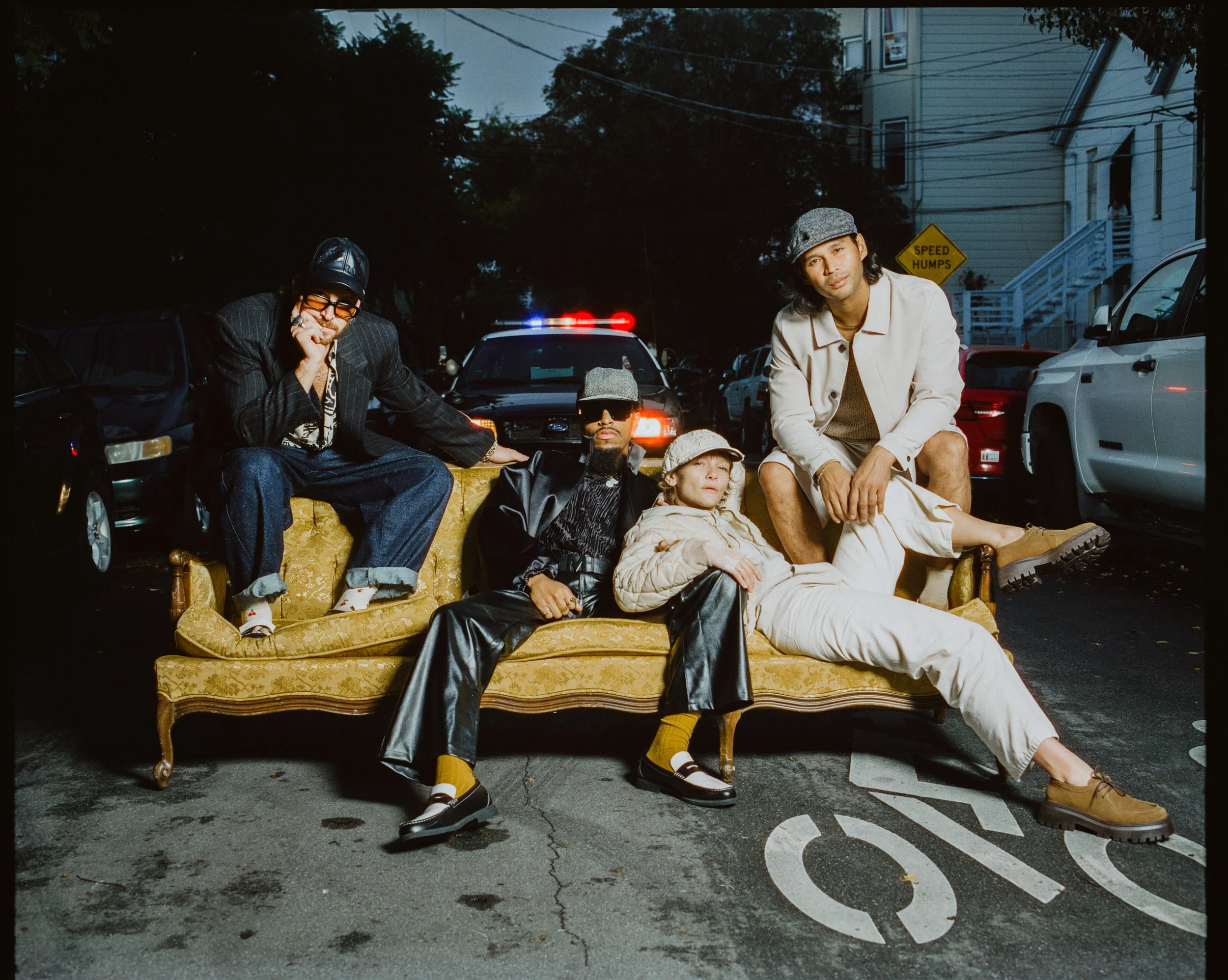 A group sitting on a vintage yellow couch outdoors at night, surrounded by parked cars and houses, with a police car with flashing lights in the background.