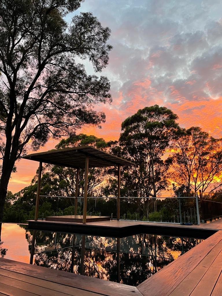 A reflective pool surrounded by a wooden deck, with trees and a sunset sky in the background.