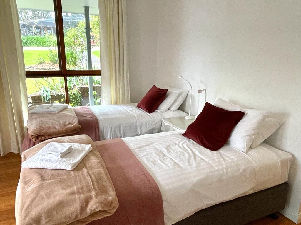 Cozy bedroom with two single beds, white linens, maroon pillows, brown blankets, a bedside table with reading lamps, and a large window with garden view.