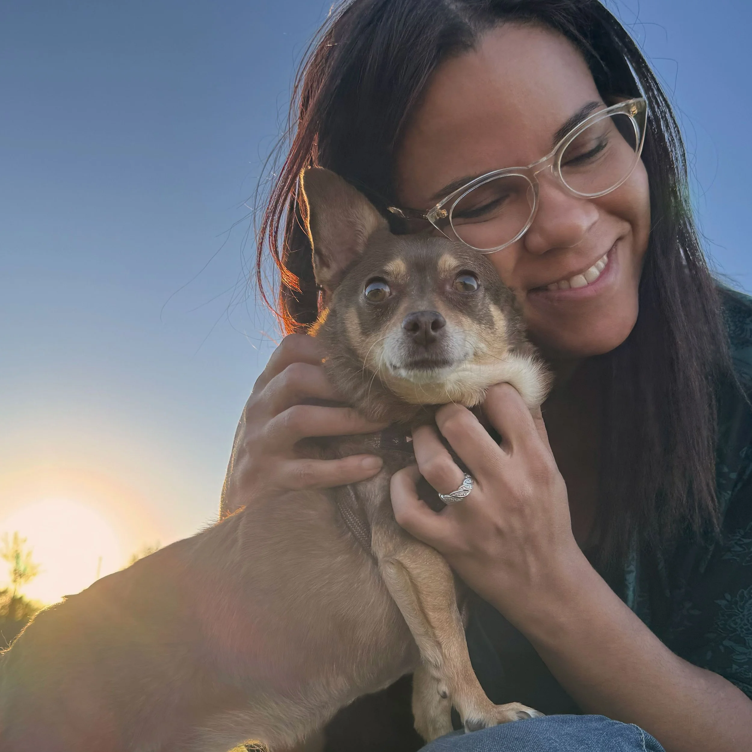 A woman with glasses smiling and holding a small brown dog with a tan face during sunset, with the sun setting in the background and trees visible.