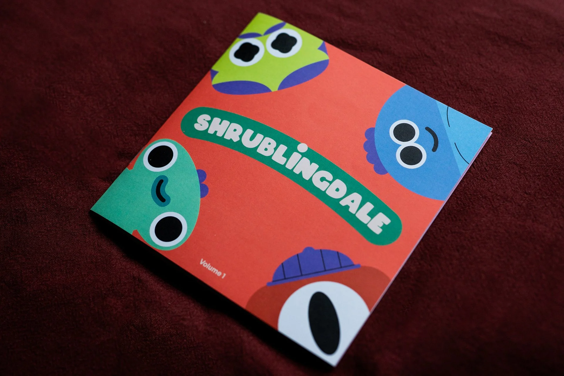 A photo of the Shrublingdale front page, which is square, orange with a logo in the centre and several monsters popping in from the sides.