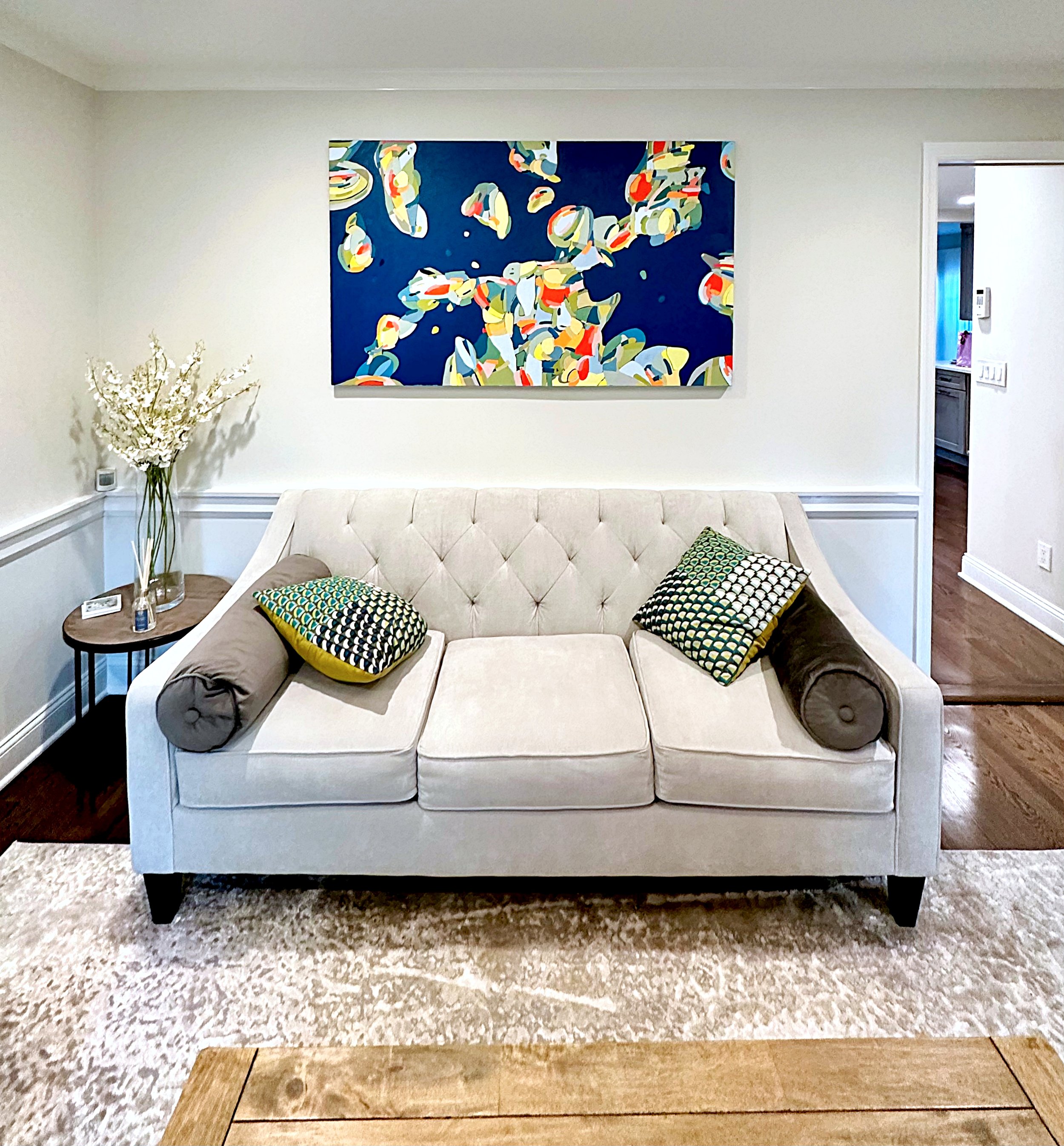 Living room with a beige tufted sofa, two patterned pillows, a round black side table with a tall vase of white flowers, a large colorful abstract painting on the wall, and a doorway leading to a hallway with hardwood floors.
