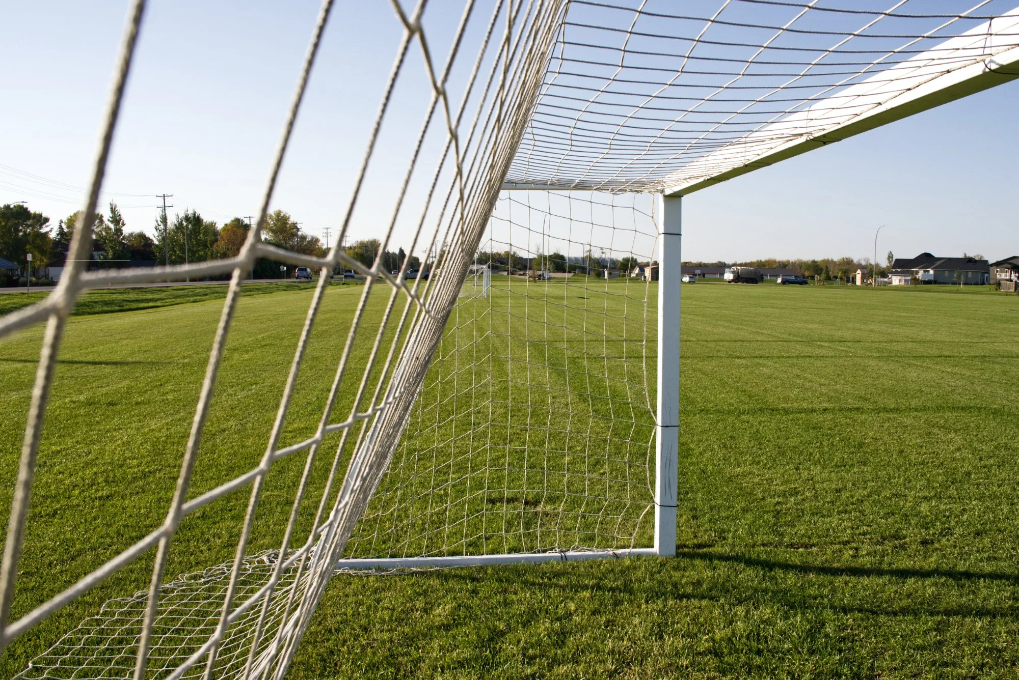 Close-up view of a soccer goal net on a grassy field with suburban houses in the background under a clear sky.