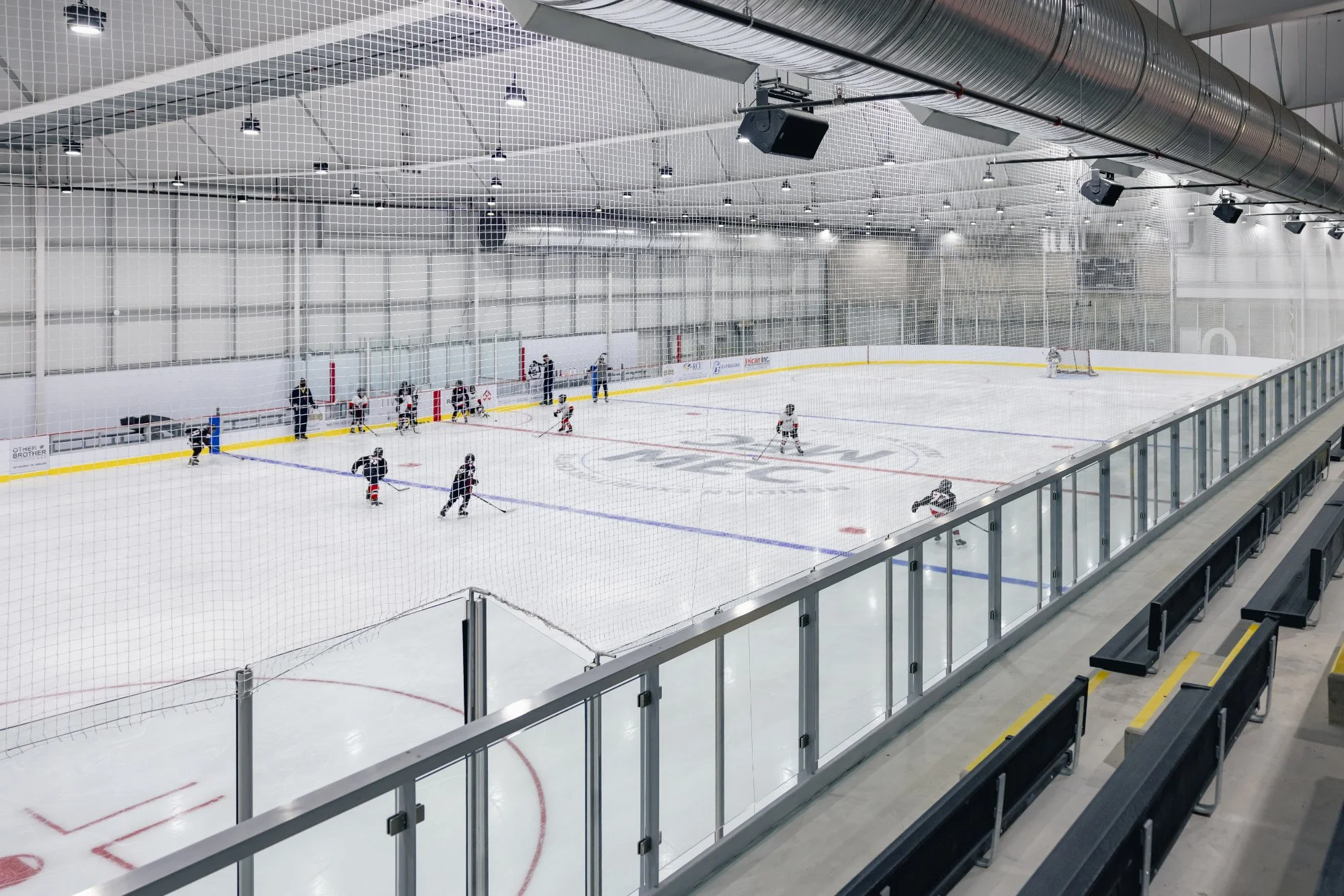 Indoor ice hockey rink with players practicing on the ice, surrounded by glass barriers and spectator benches.