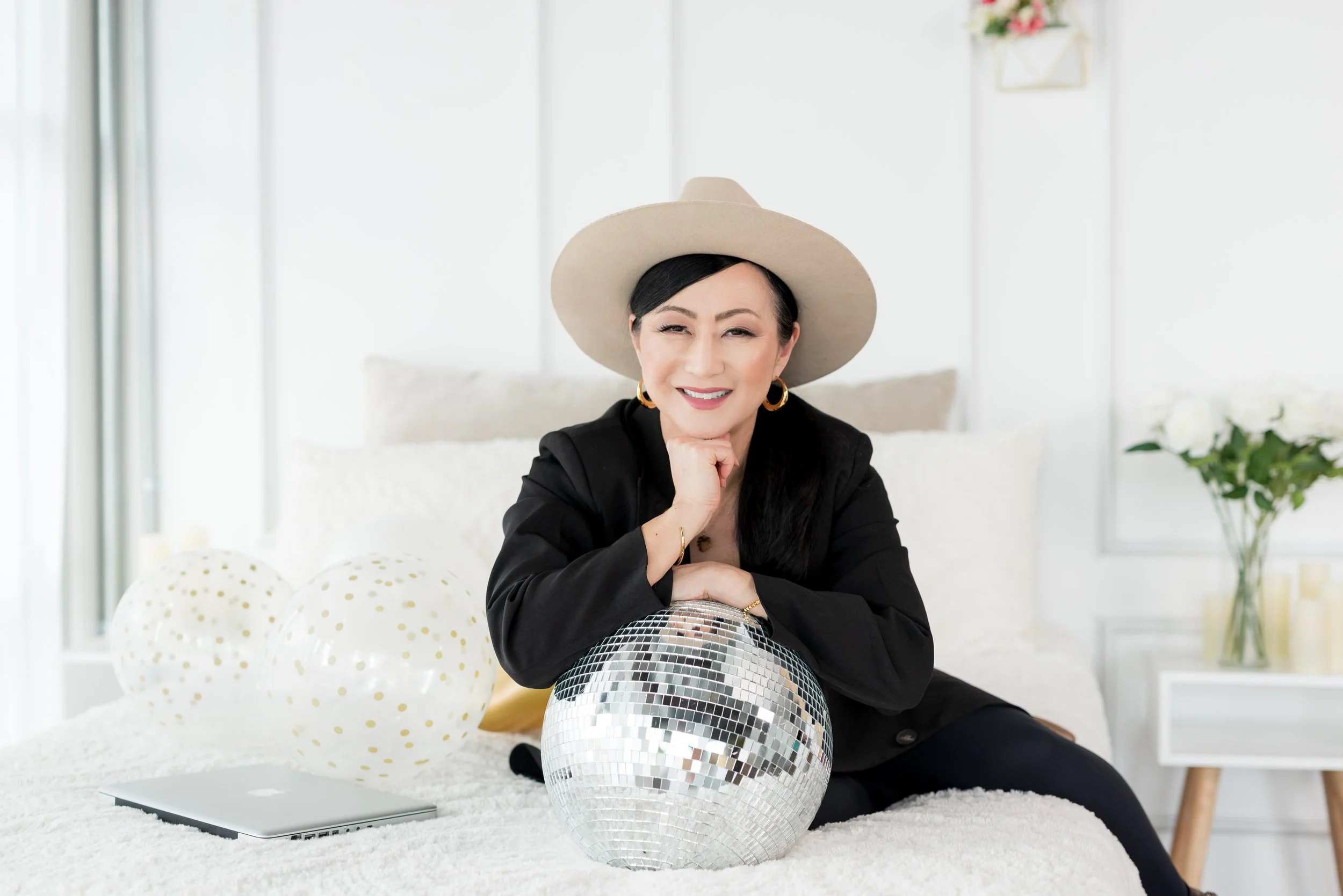 Female founder in a hat leaning over a disco ball with a laptop beside her during a studio brand photoshoot in San Diego