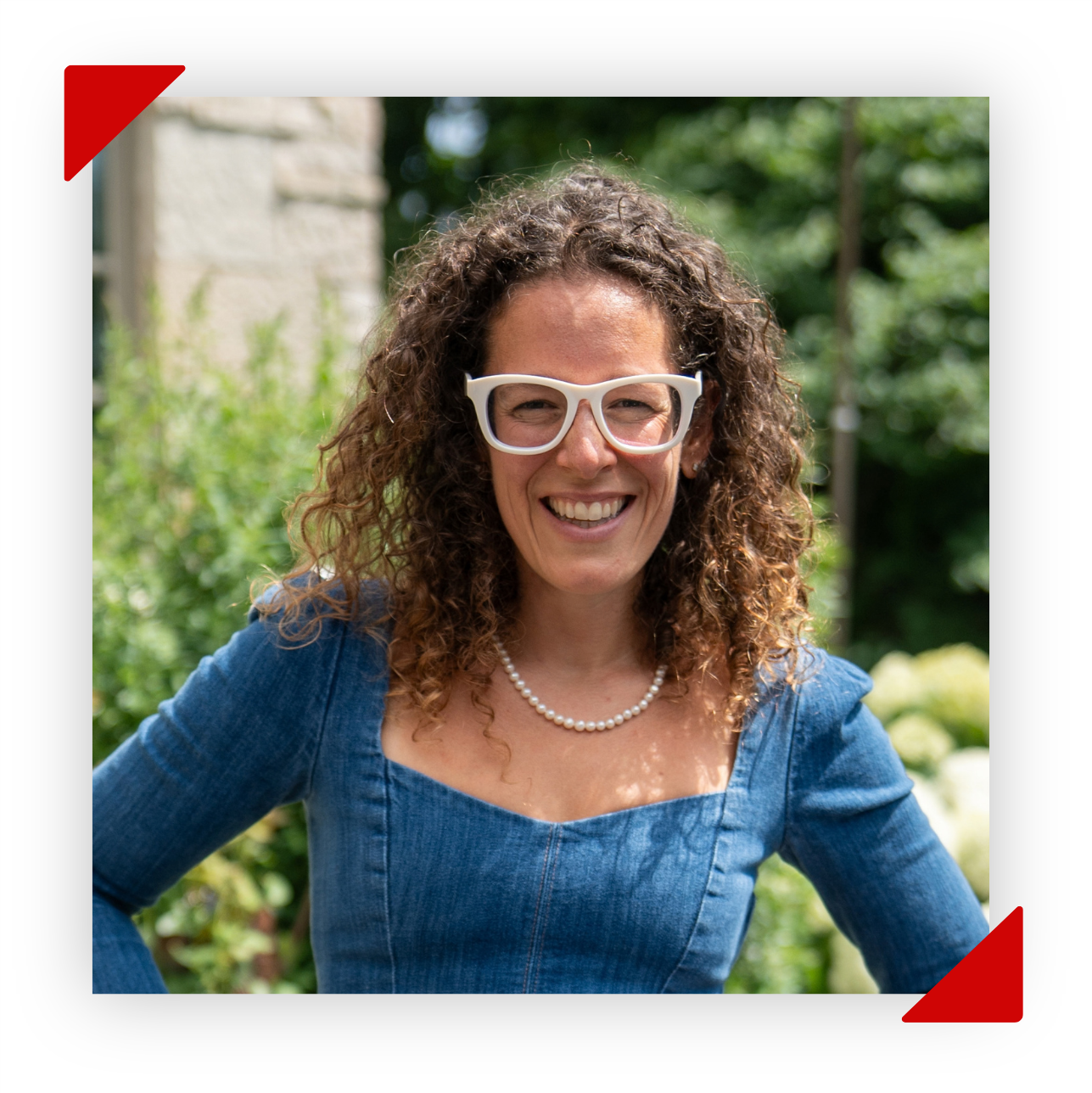 A woman with curly hair wearing white glasses, pearl necklace, and a navy blue floral dress, smiling broadly.
